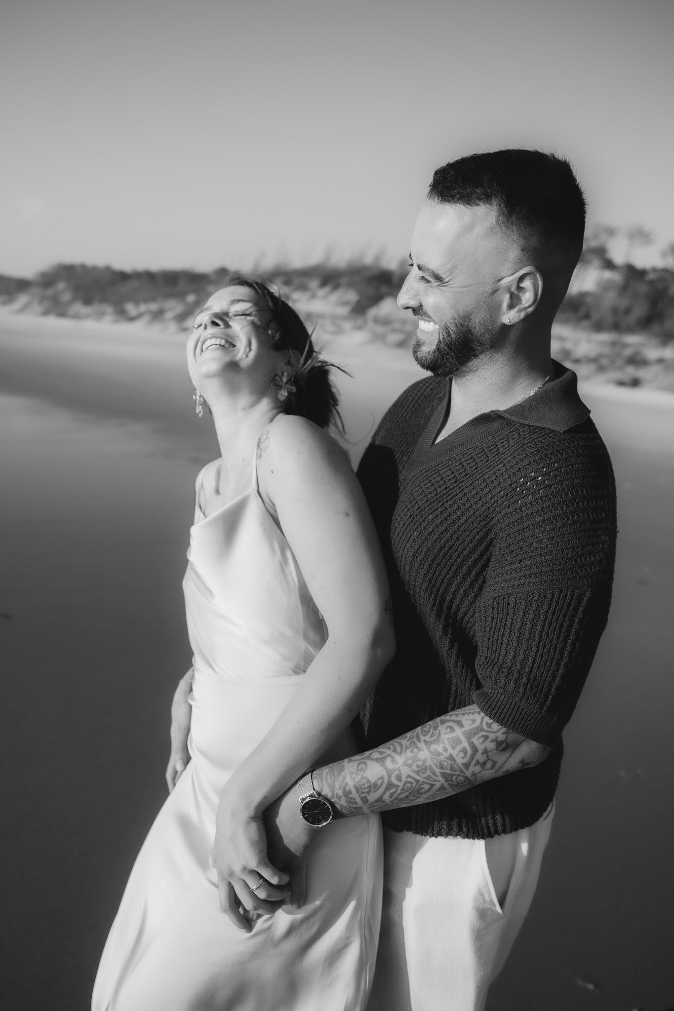 Romantic couple in white dress and dark sweater posing at beach during sunset.