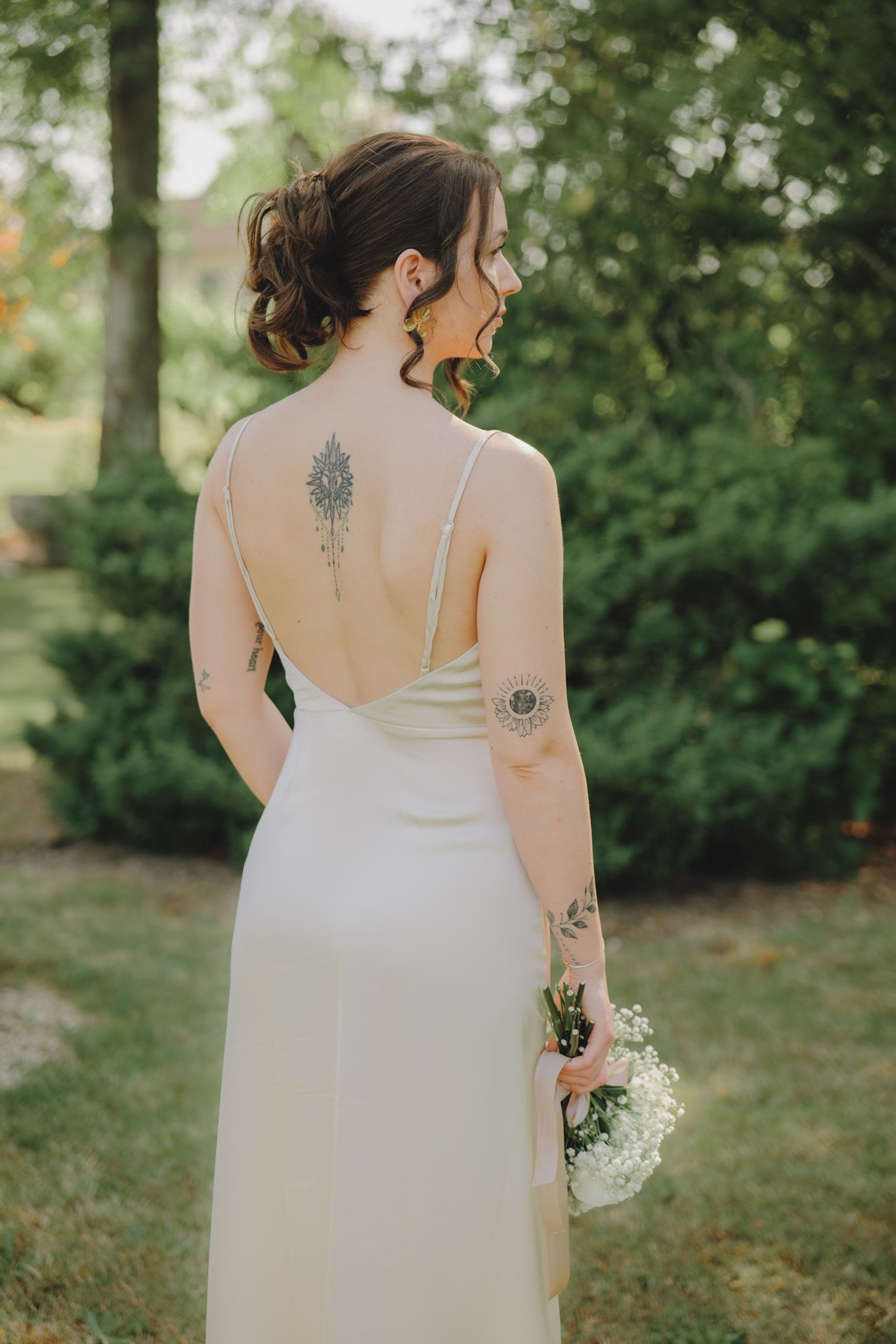 Bride in sleeveless white gown displays artistic tree tattoo on back in outdoor garden setting.