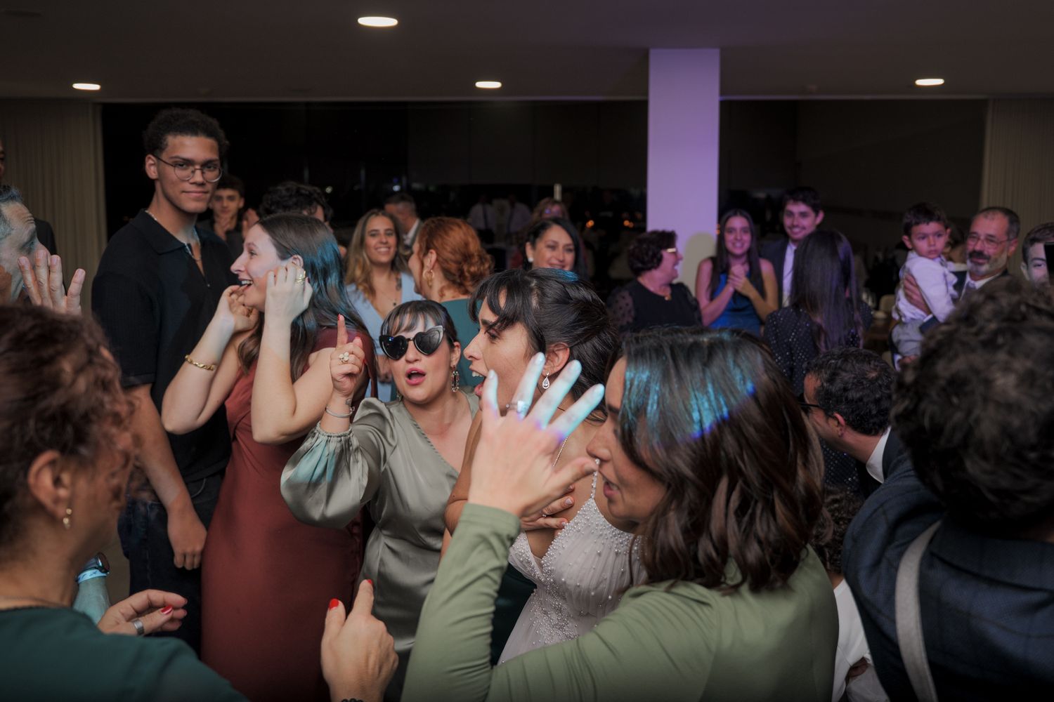 Wedding guests dance and celebrate on a crowded dance floor under purple lighting.