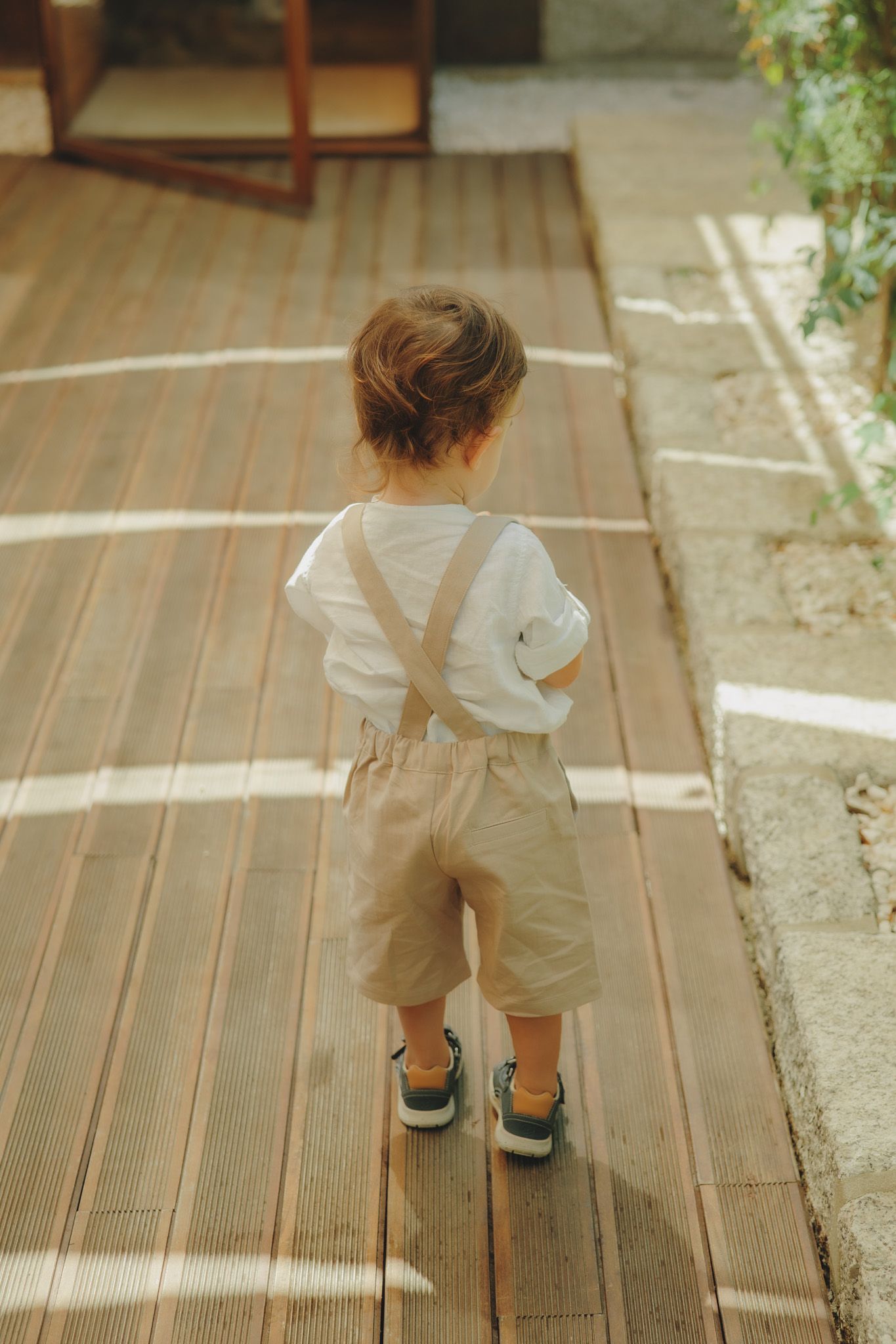 Small child in overalls walks along a wooden deck in sunlight.