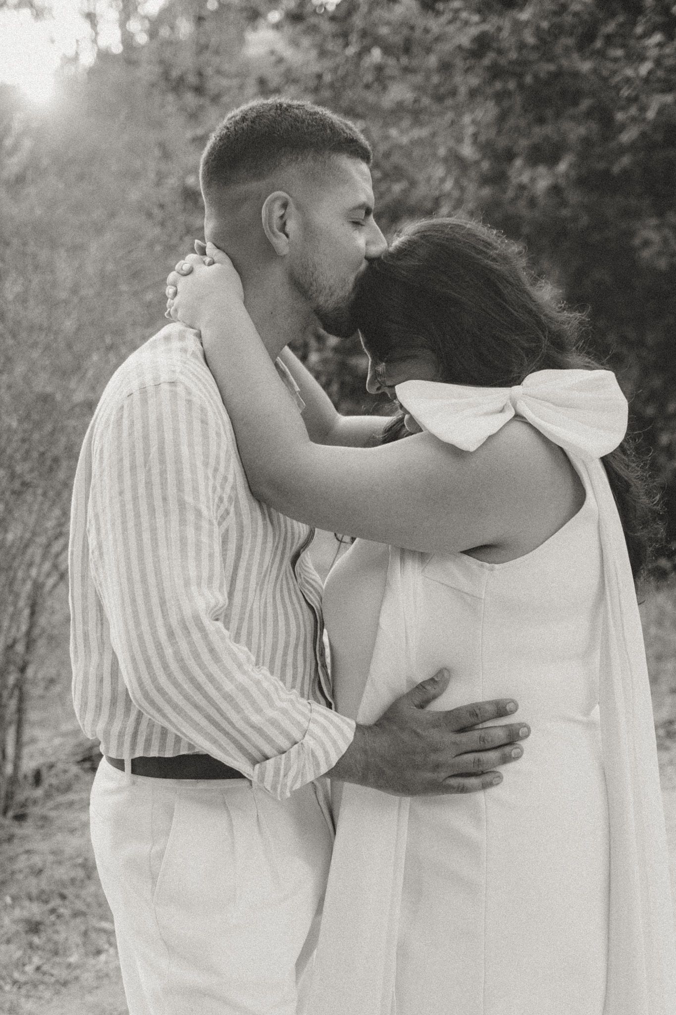A couple embraces tenderly in white outfits against a natural backdrop.