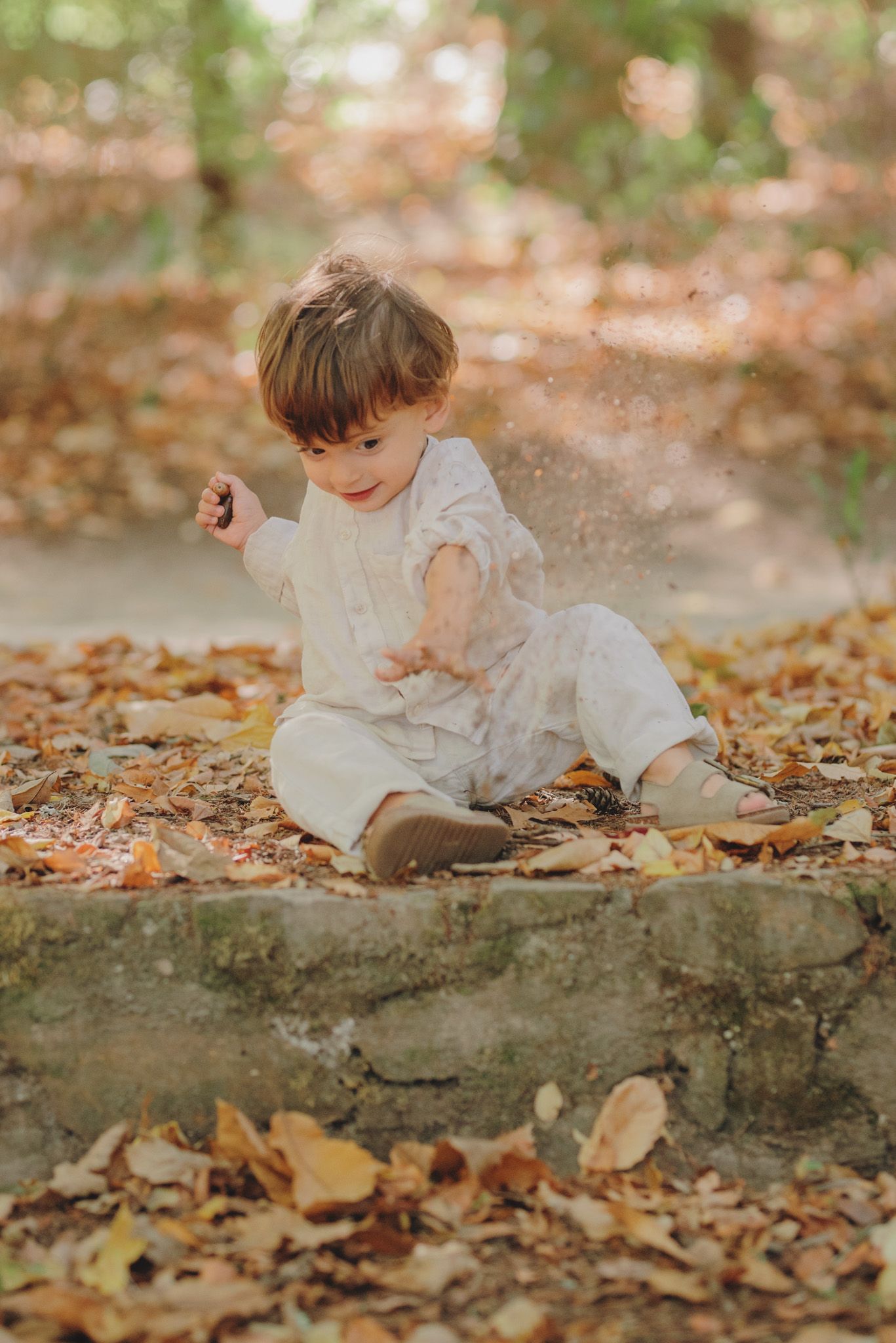 A small child in white clothing sits playfully among fallen autumn leaves.