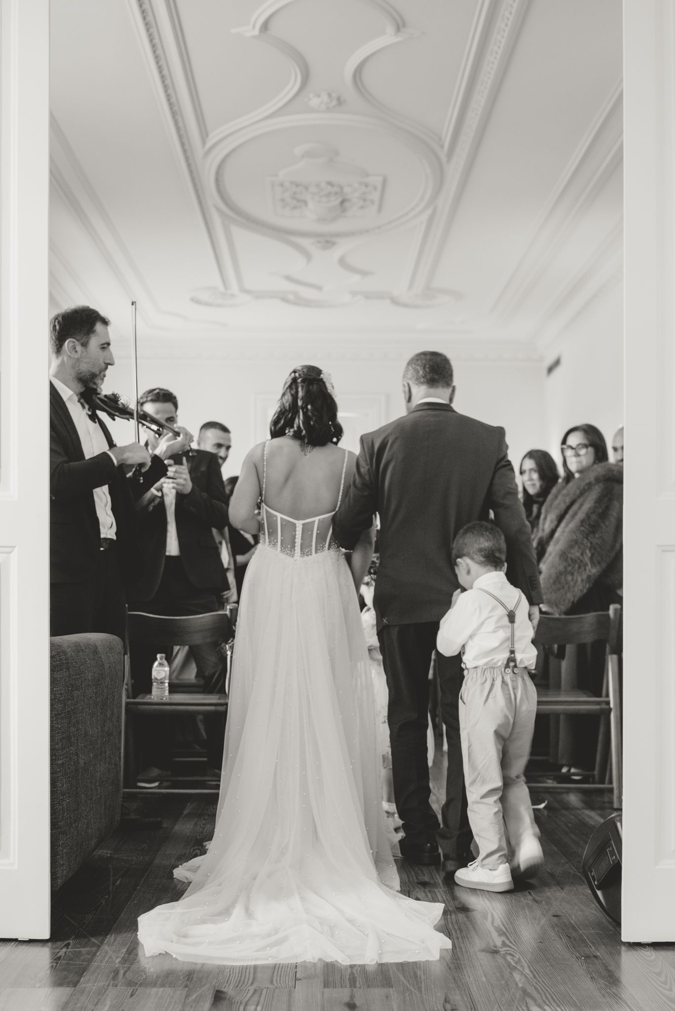 Wedding guests gather in an elegant room with ornate white ceiling details and hardwood floors during a ceremony.
