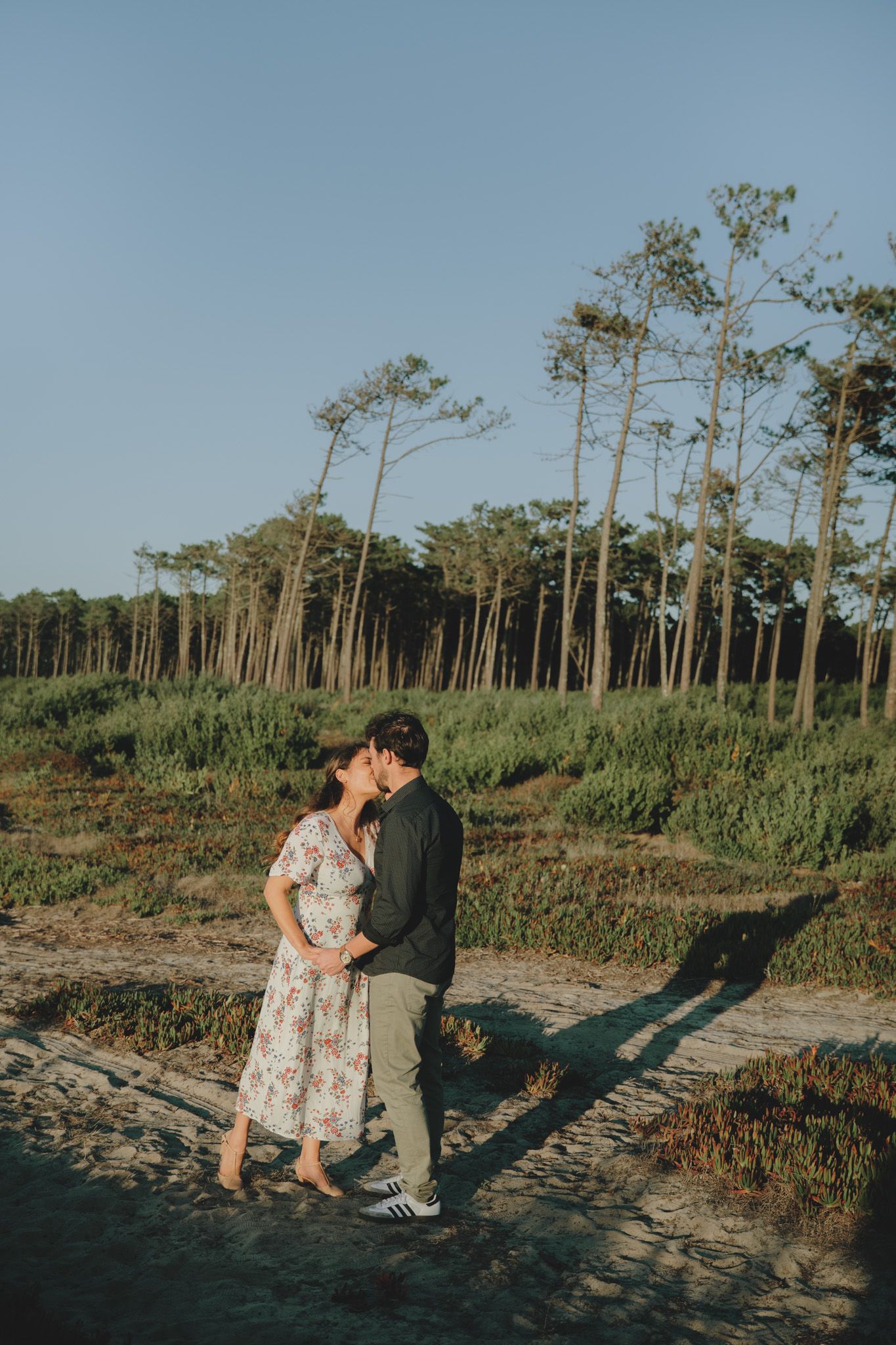 A couple embraces on a beach path with tall pine trees in the background.