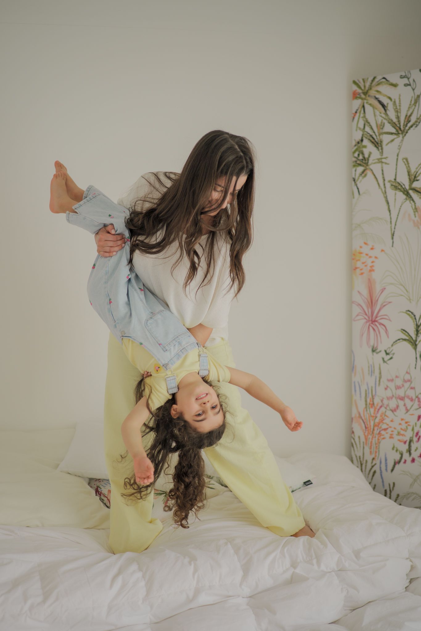 A woman playfully holds a laughing child upside down on a white bed near a floral wall art.