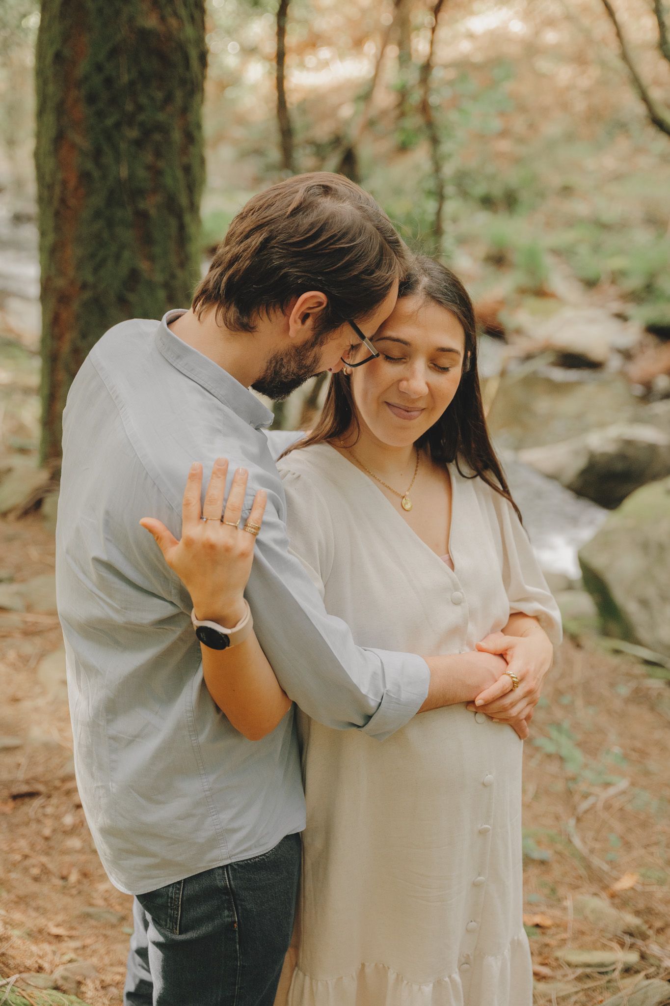 A couple embraces in a forest setting while showing off a ring.