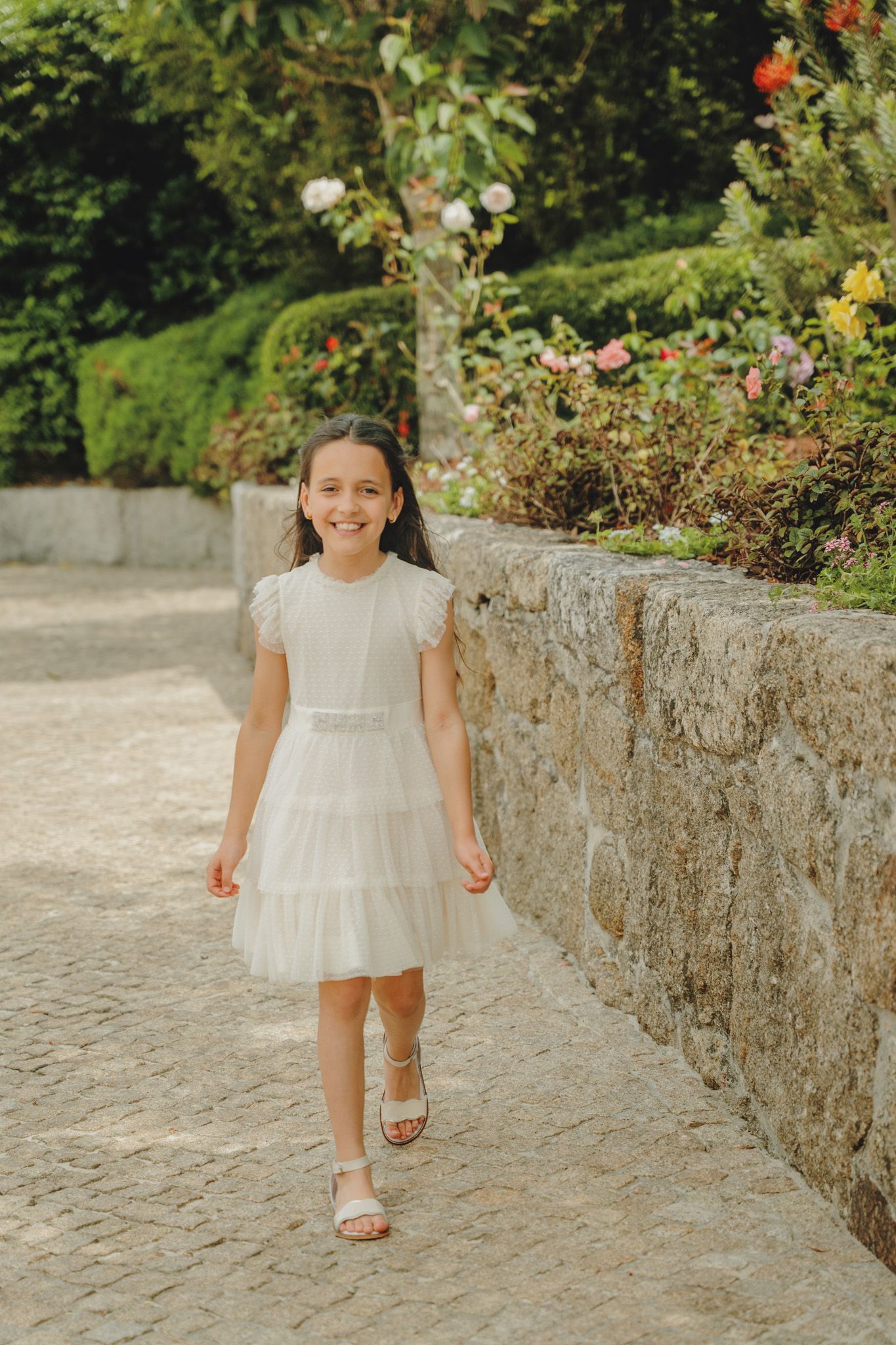 A little girl in a white dress walks along a stone path with flowers and greenery in the background.