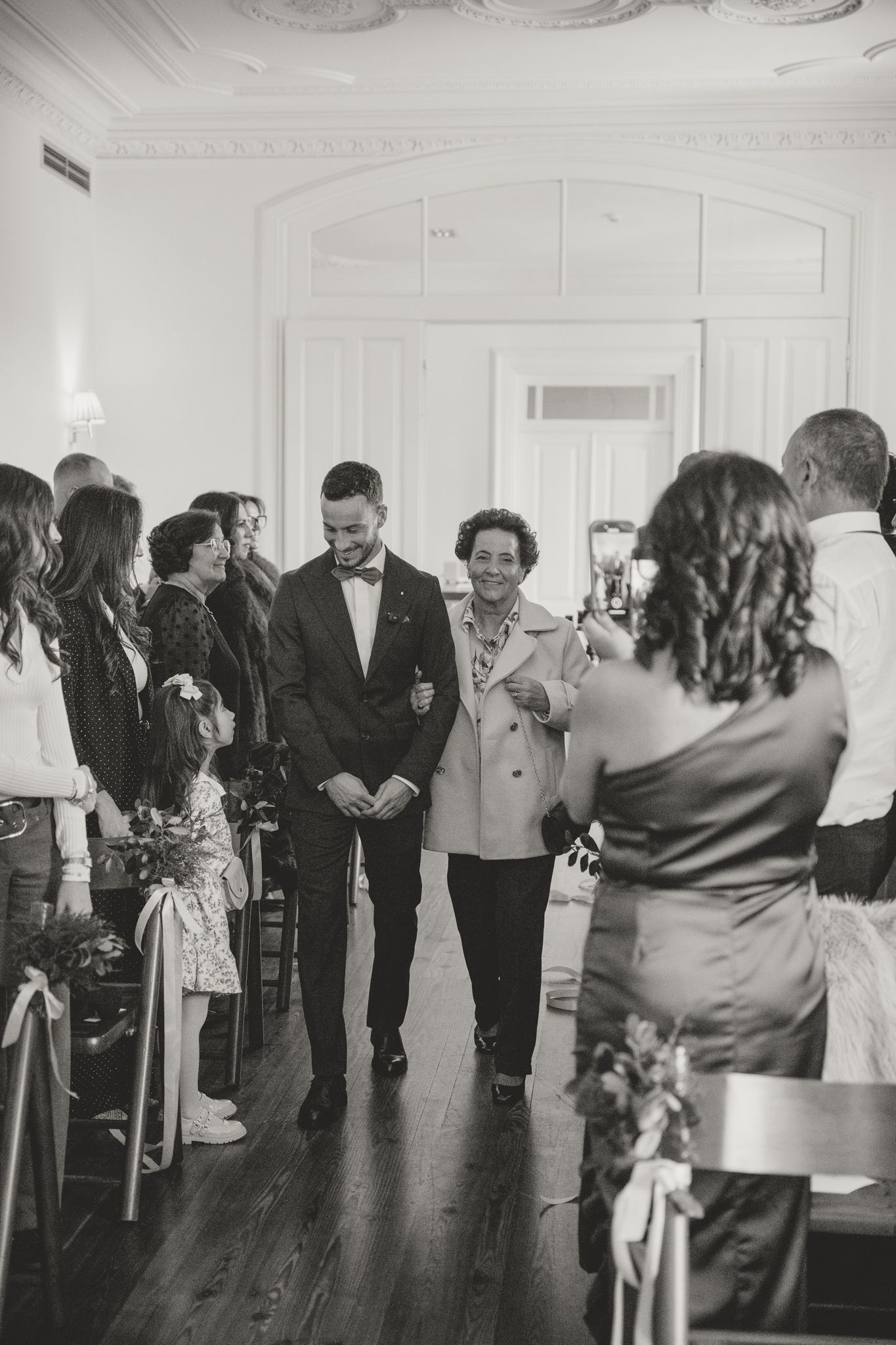 Guests socialize in an elegant white-walled room with hardwood floors during a celebration.