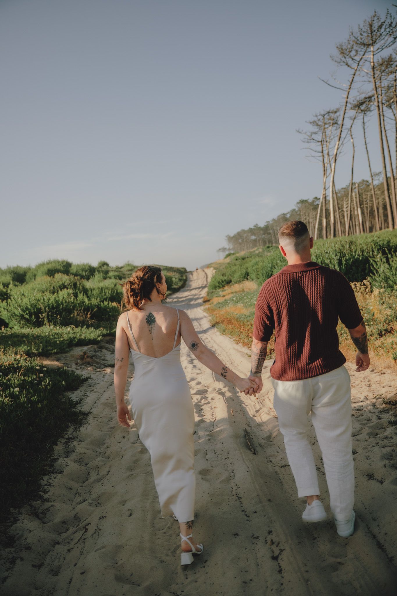 People strolling down a sandy beach path surrounded by pine trees and coastal vegetation.
