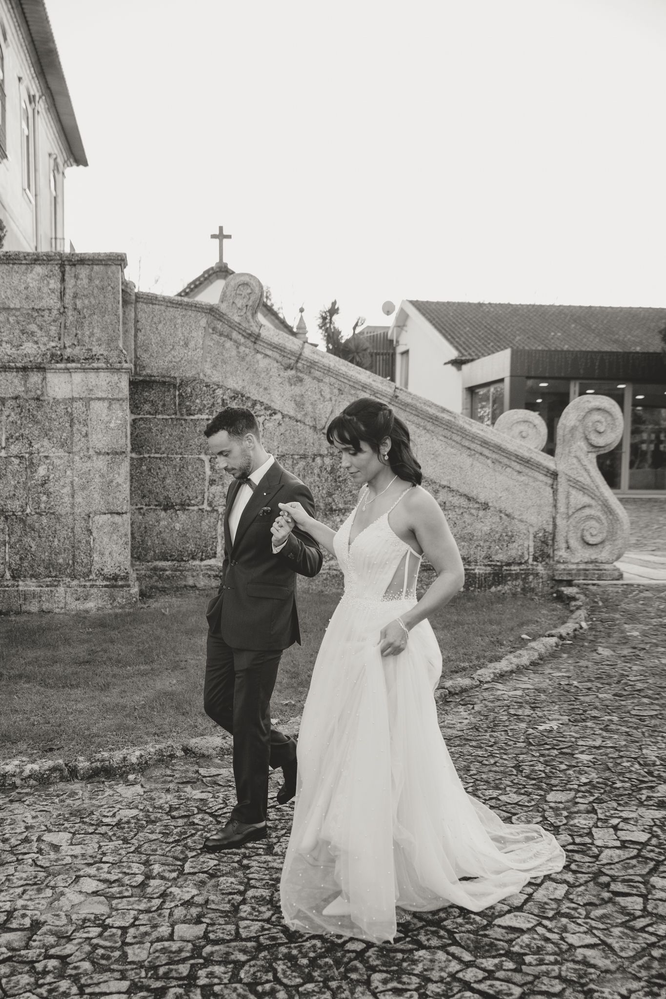 Wedding couple walking on cobblestone path outside historic stone building.