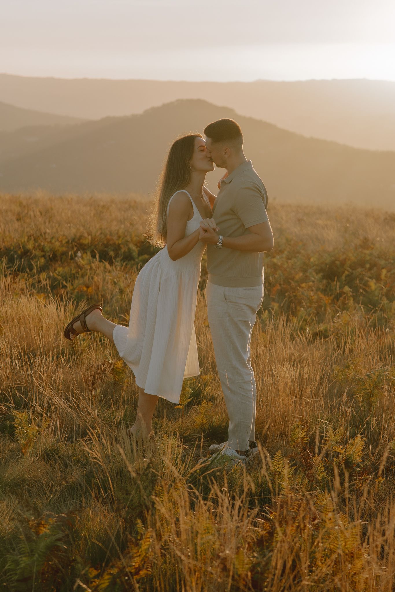 A couple embraces in a golden field at sunset with mountains in the background.