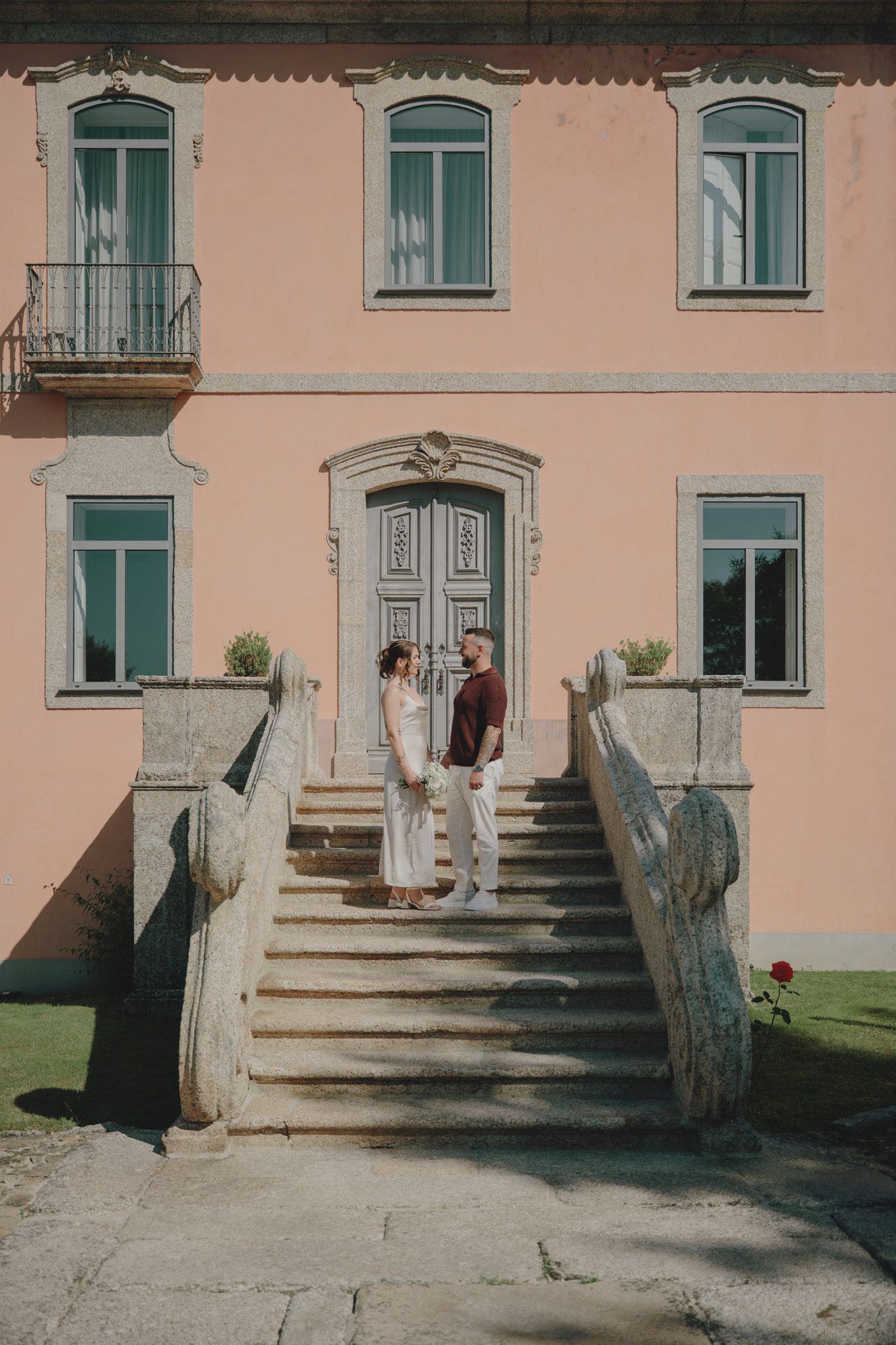 A couple poses on stone steps in front of a pink building with white trim and arched windows.