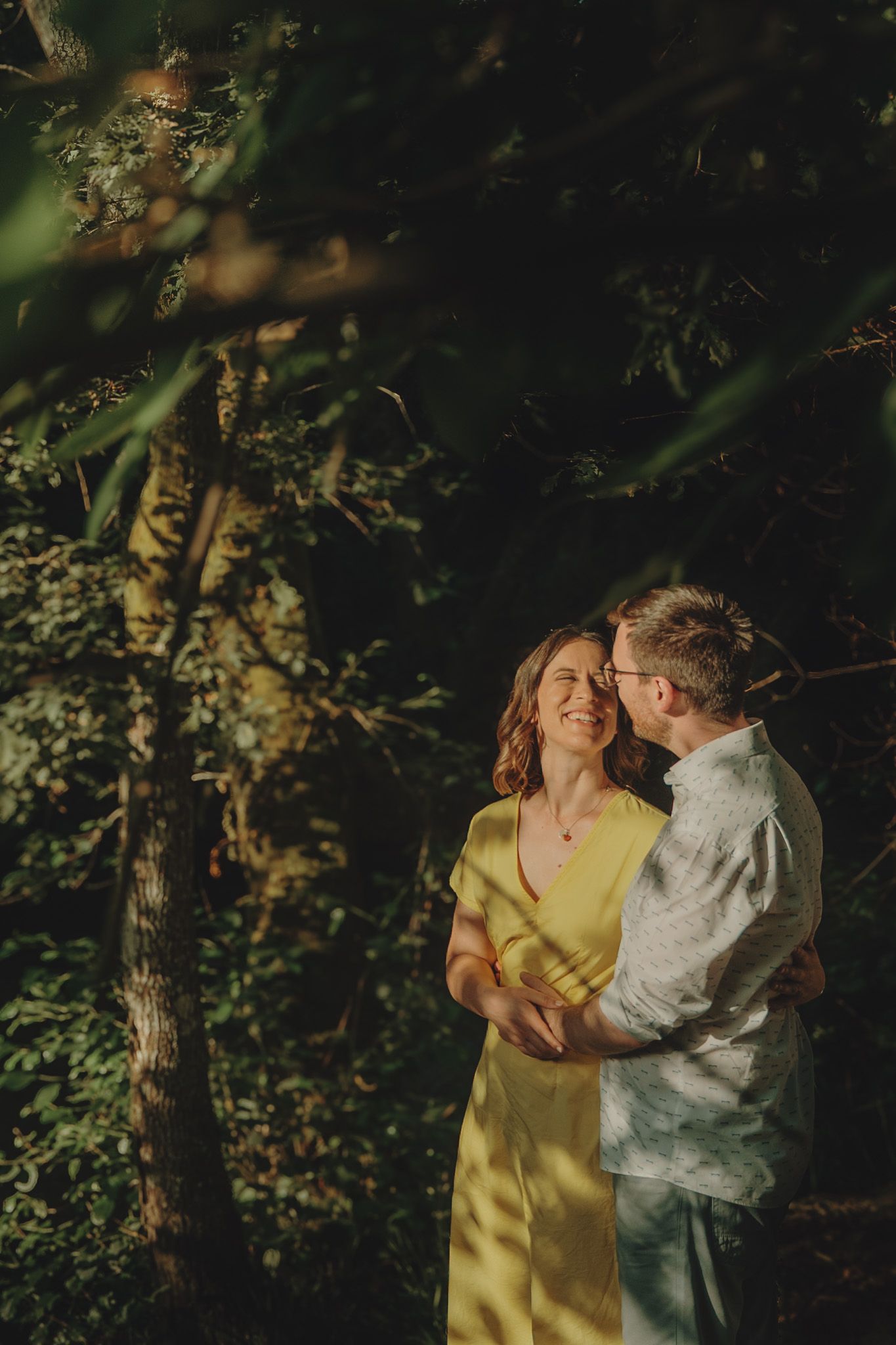 A couple embraces in golden sunlight among trees wearing yellow and white clothing.