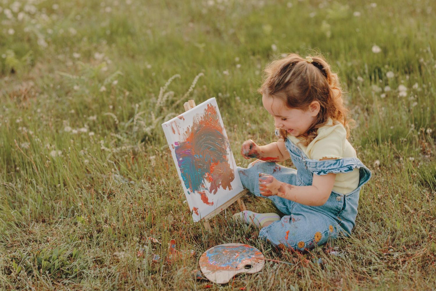Small artist in denim overalls focuses intently on their painting while sitting in a wildflower field.