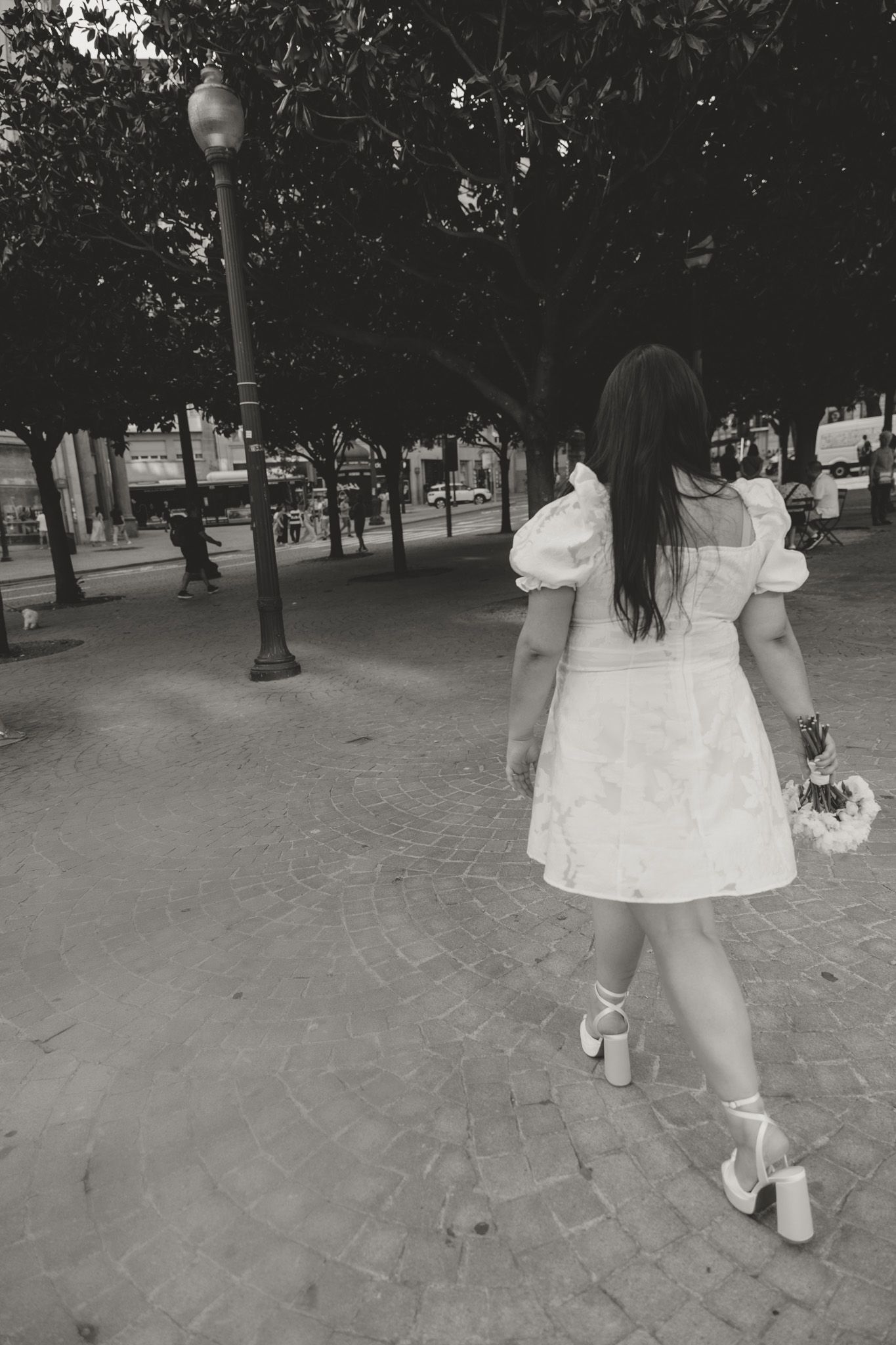 A person in a white dress walking away on a tree-lined cobblestone path.