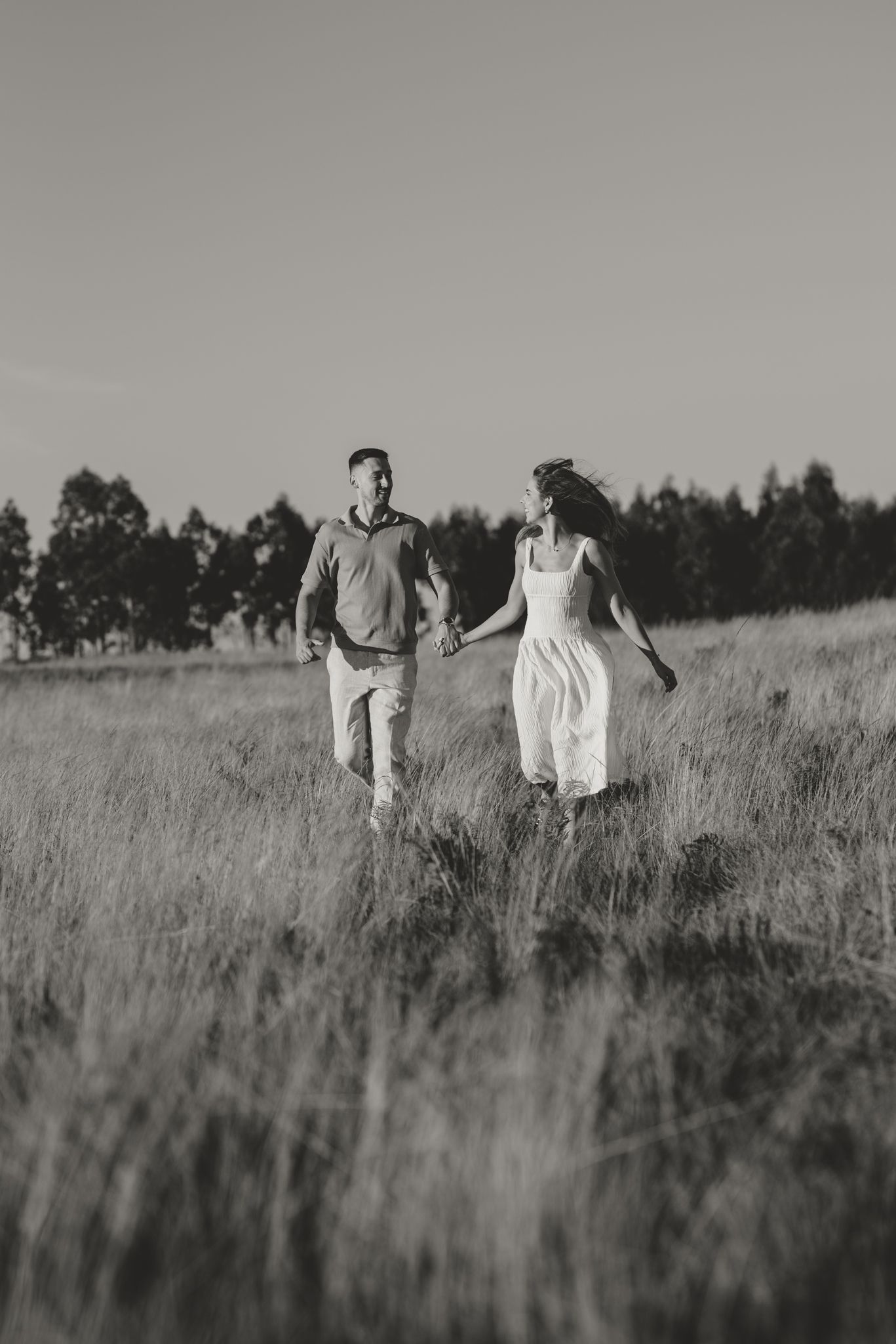 Black and white photo of two people walking through a serene meadow with trees in the background.