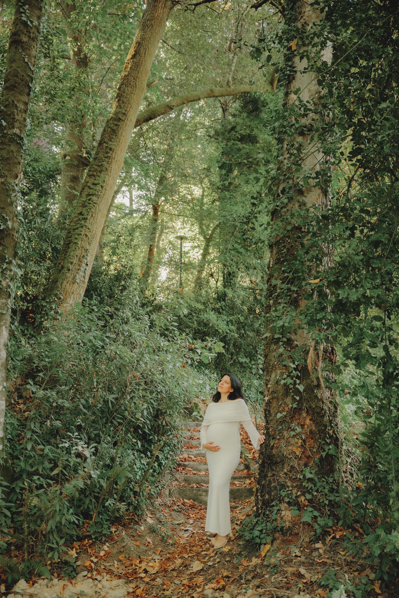 Full-length maternity portrait in a sunlit forest wearing a fitted white gown.
