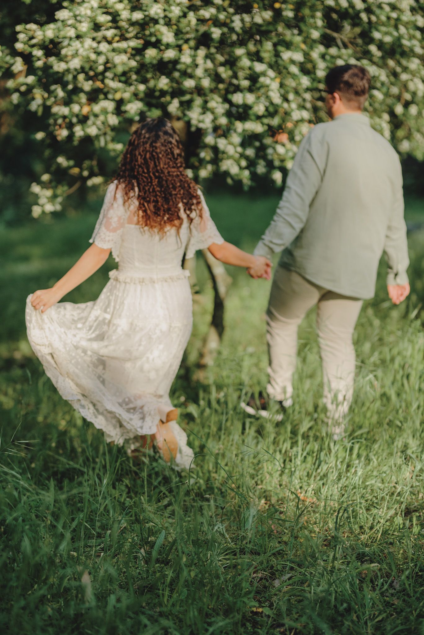 Couple walking through tall grass with flowing white dress trailing behind.