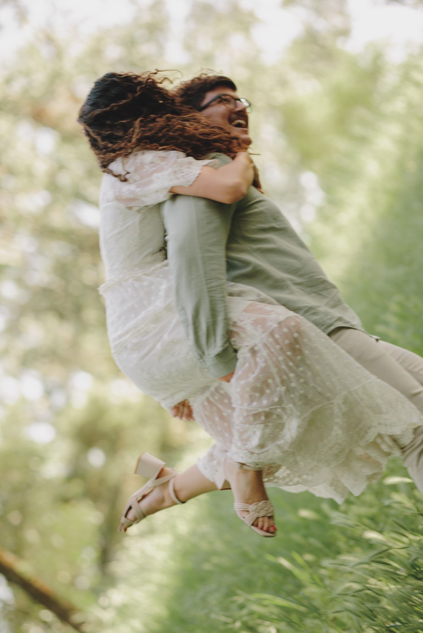 A couple in romantic motion spins together outdoors with the woman's white dress flowing in the sunlight.