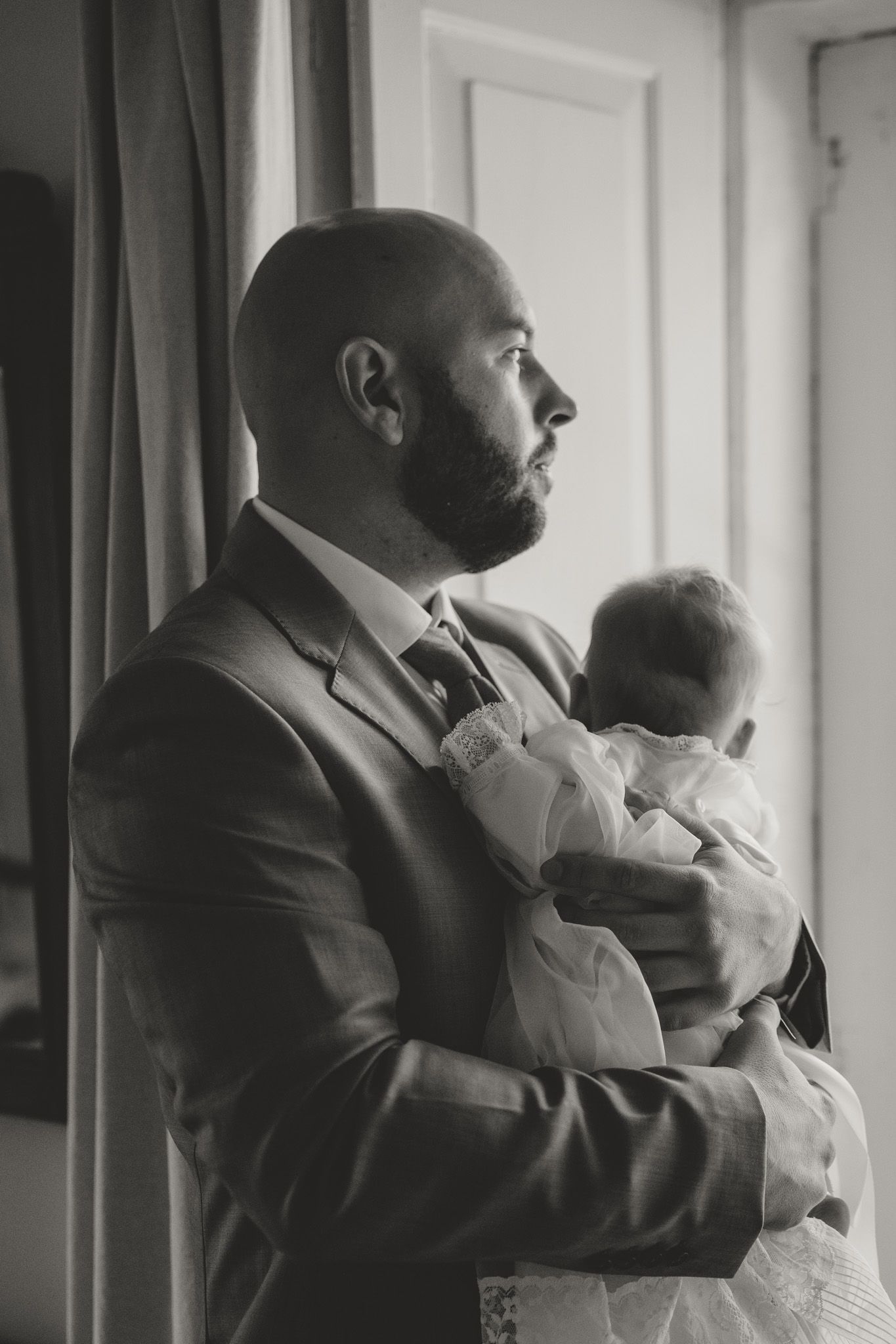 A black and white photograph of a suited man in profile holding a small infant by a window.