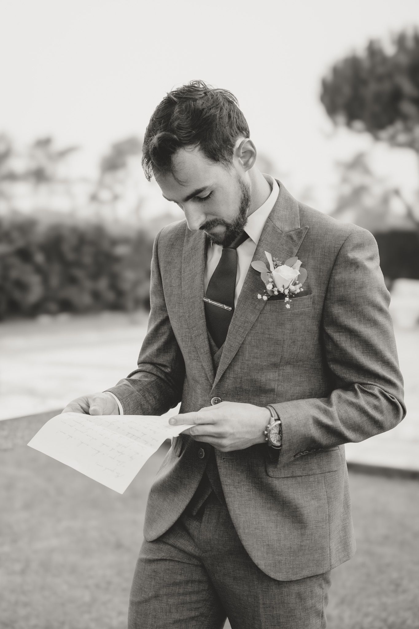 Person in grey suit reading letter outdoors during wedding ceremony.