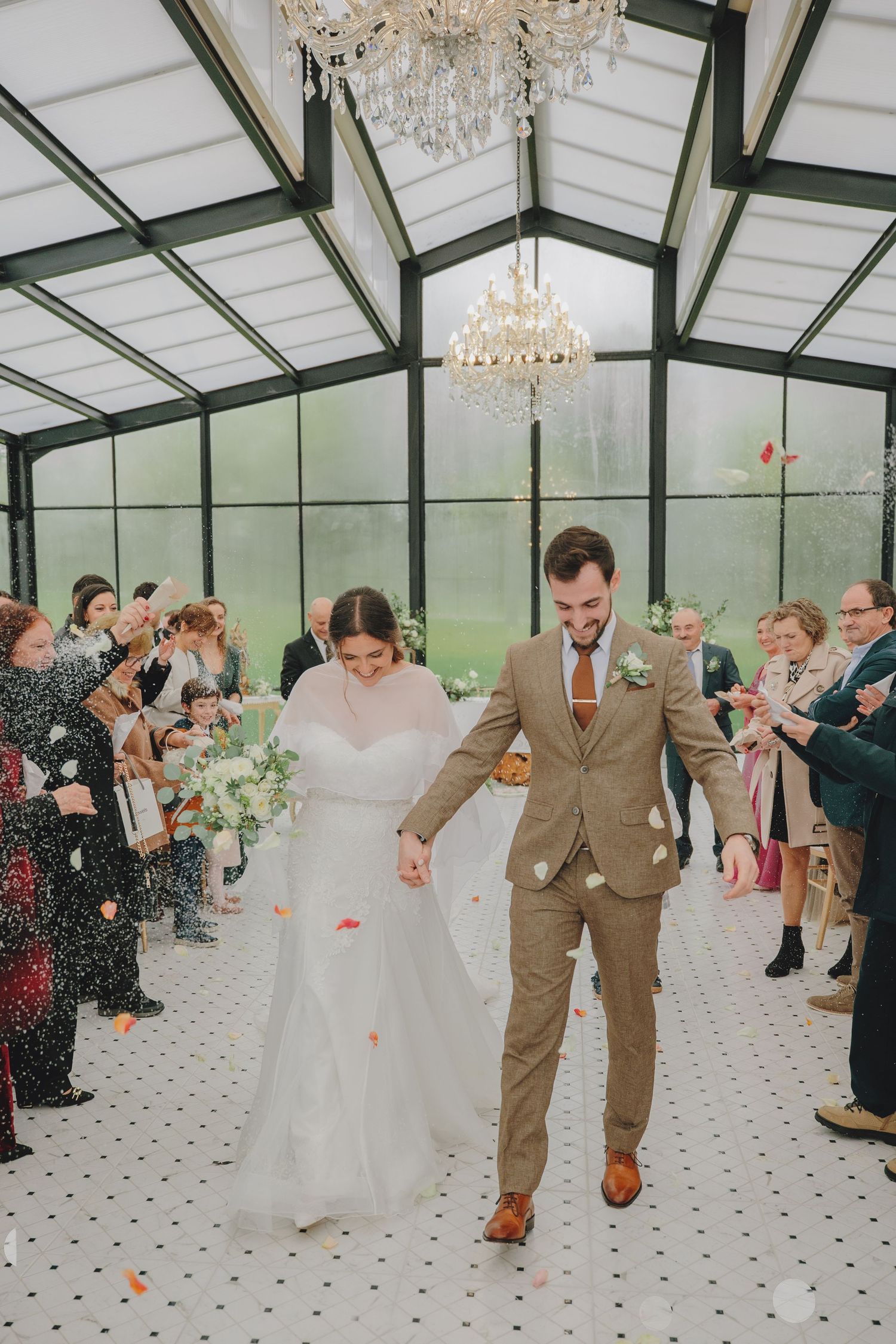 Newlyweds walk down the aisle in a greenhouse venue while guests shower them with petals.