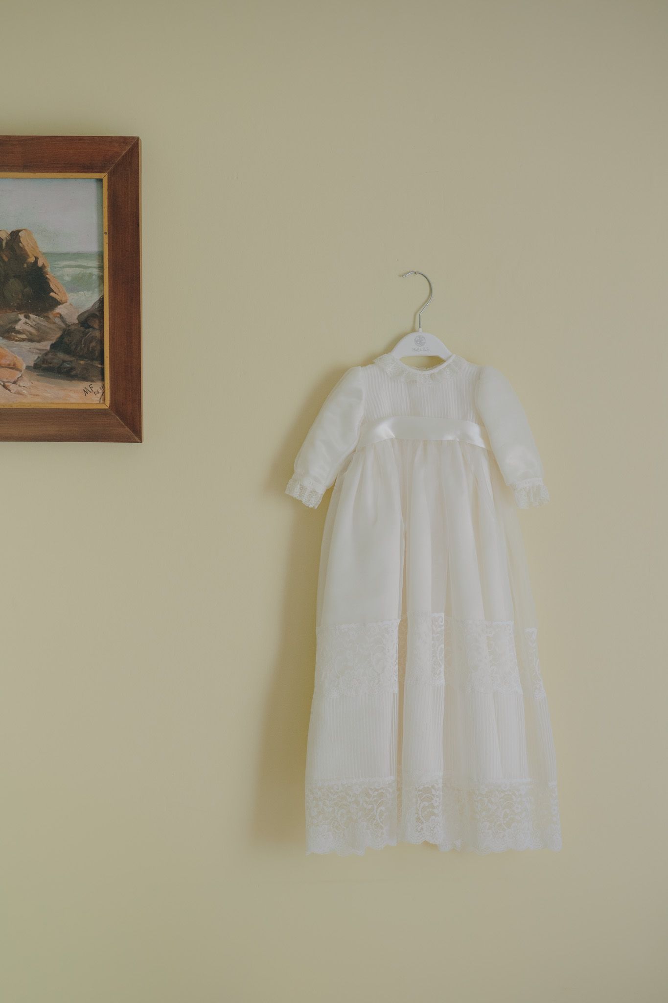 White lace christening gown hanging on display against a beige wall.