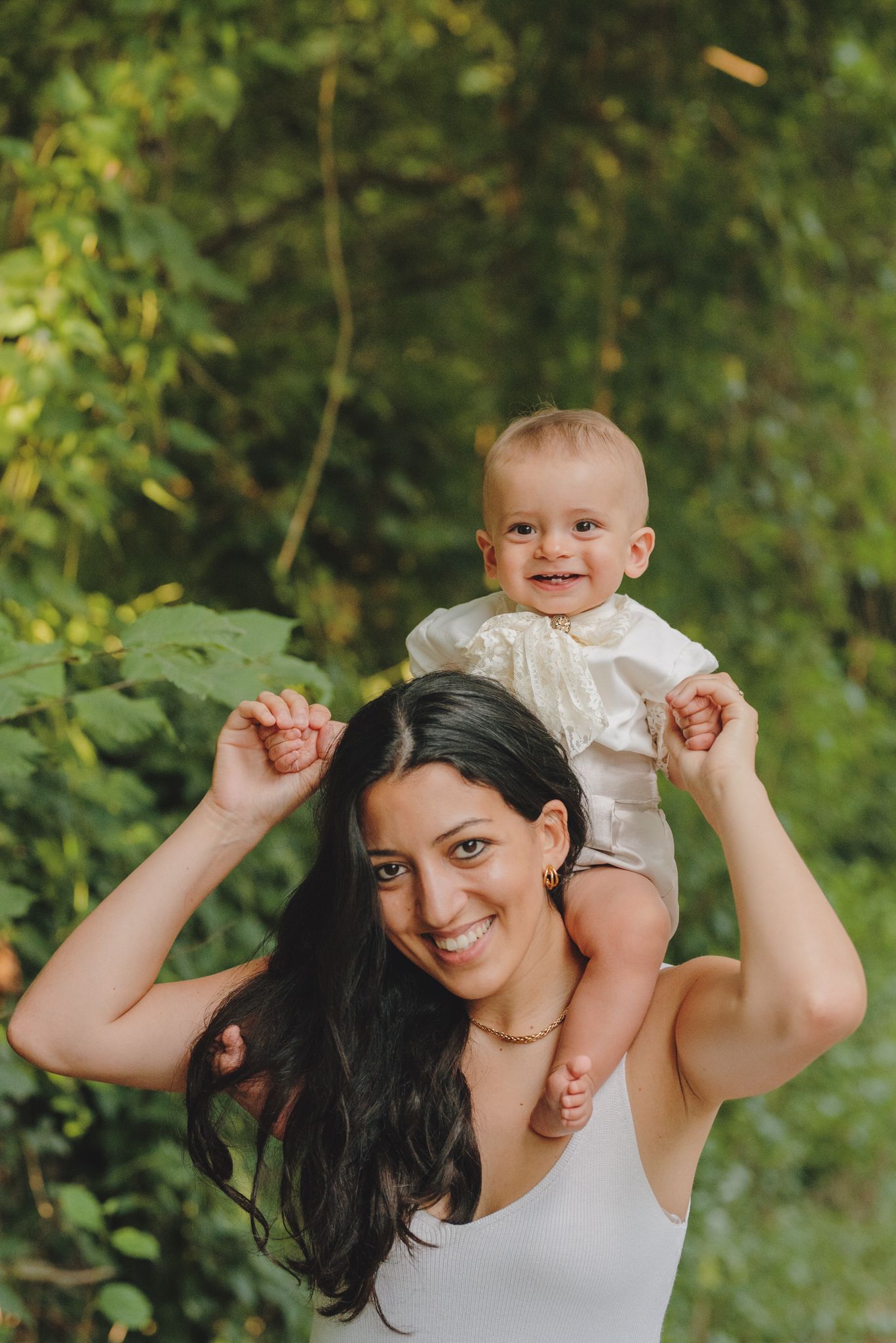 A joyful moment outdoors as a baby rides on shoulders against a green leafy background in summer.