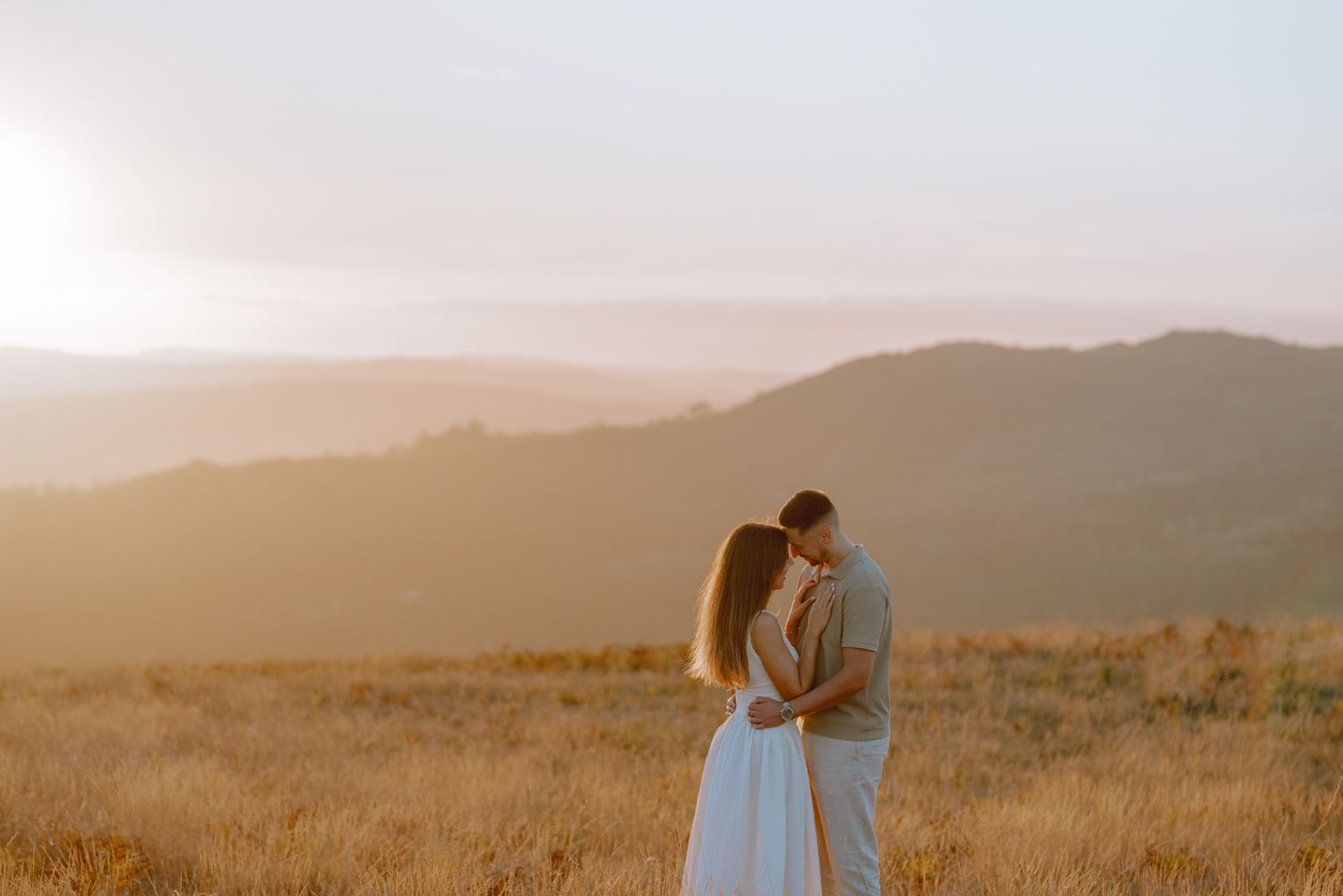 Couple embracing in a golden field during sunset with mountains in the background.