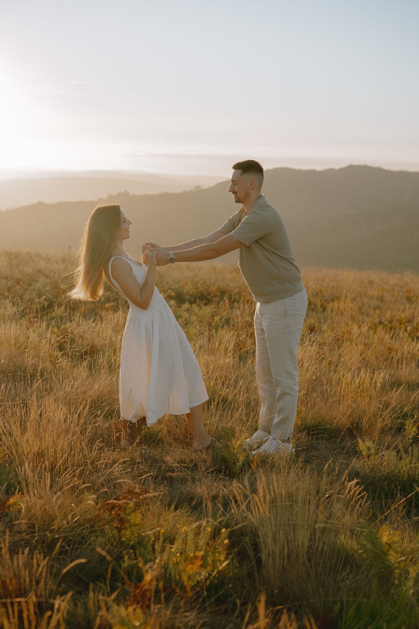 Couple dances together in a golden field during sunset with mountains in the background.