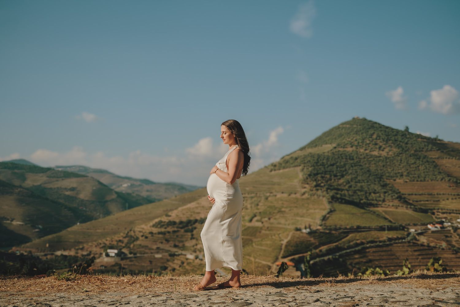 A scenic mountain landscape photoshoot featuring someone in a flowing white dress against rolling hills and blue sky.