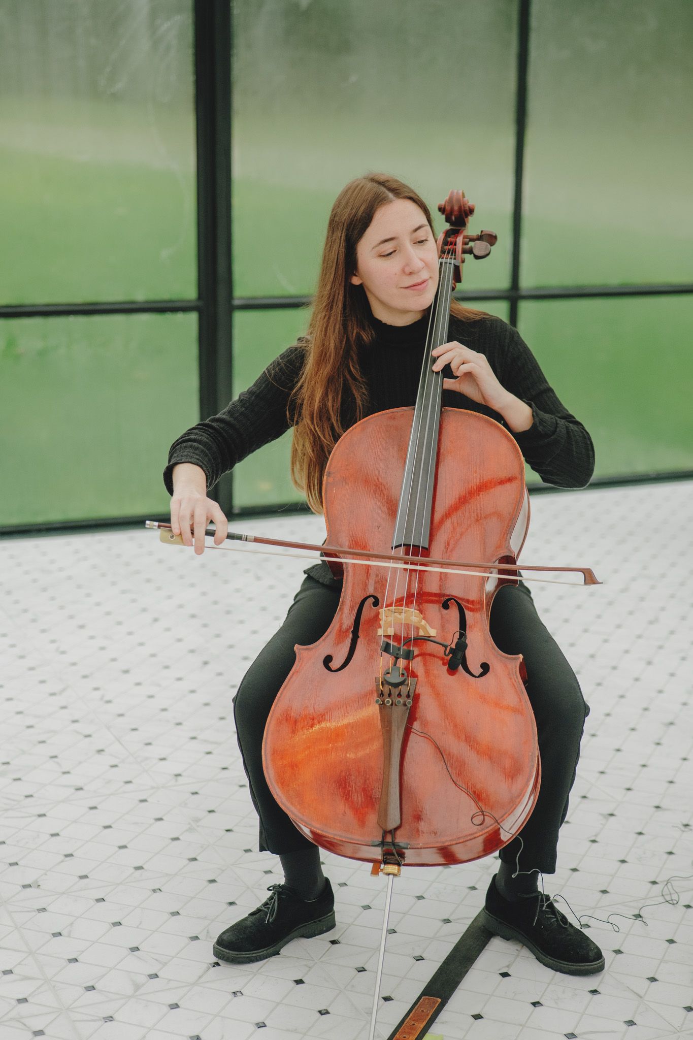 Musician plays cello in a modern glass-walled space with tiled floors.