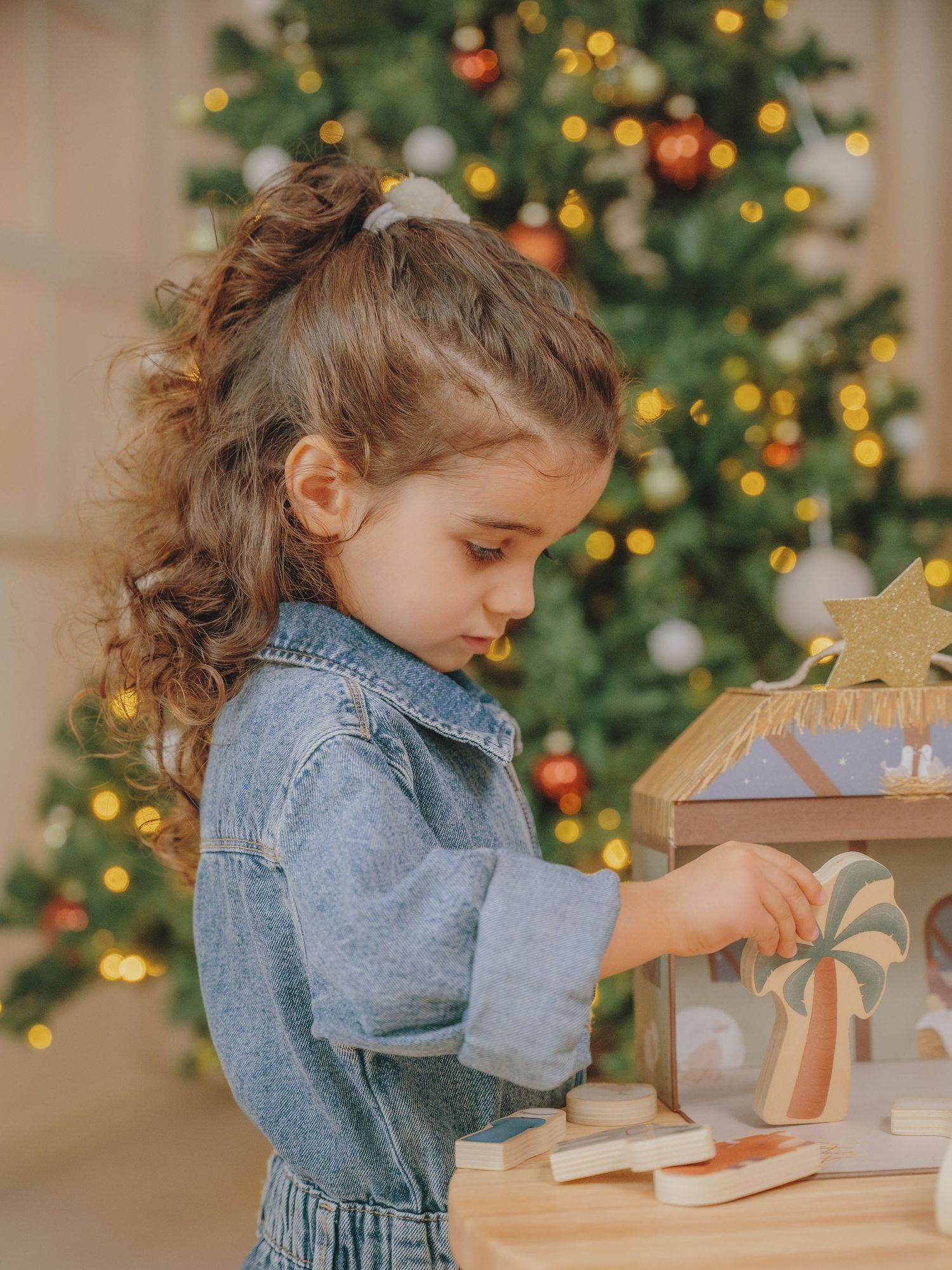 Young girl in denim dress playing with a wooden nativity set in front of a decorated Christmas tree.