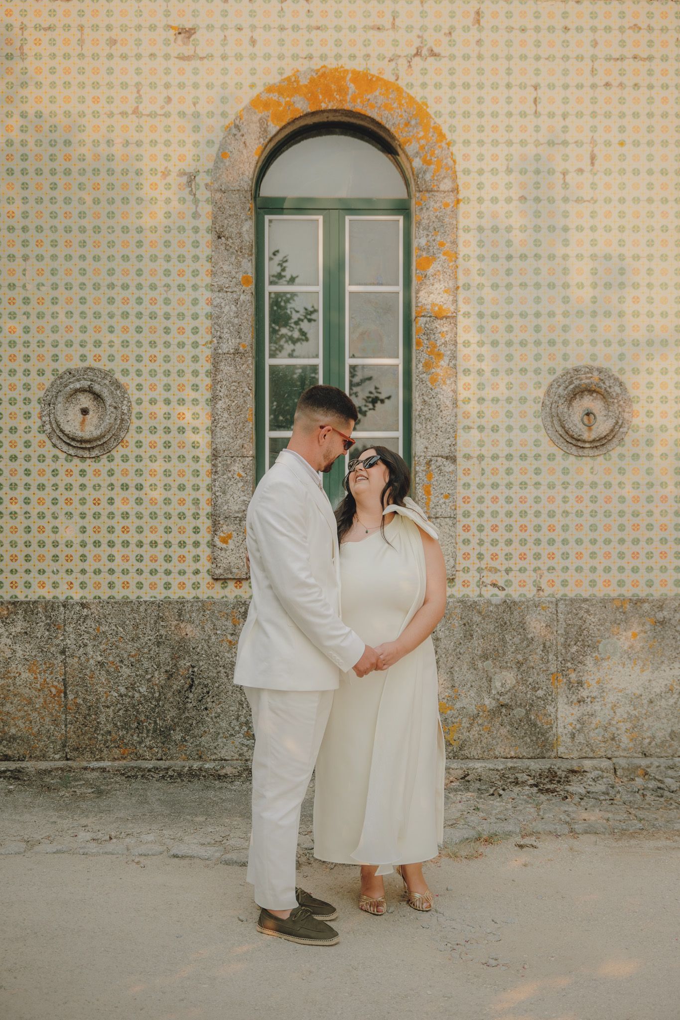 A couple in white attire poses together against a patterned tile wall.