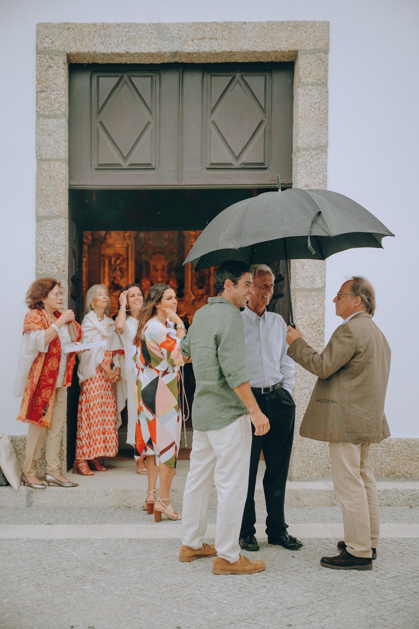 A group of people with umbrellas gathered outside a building with large wooden doors.
