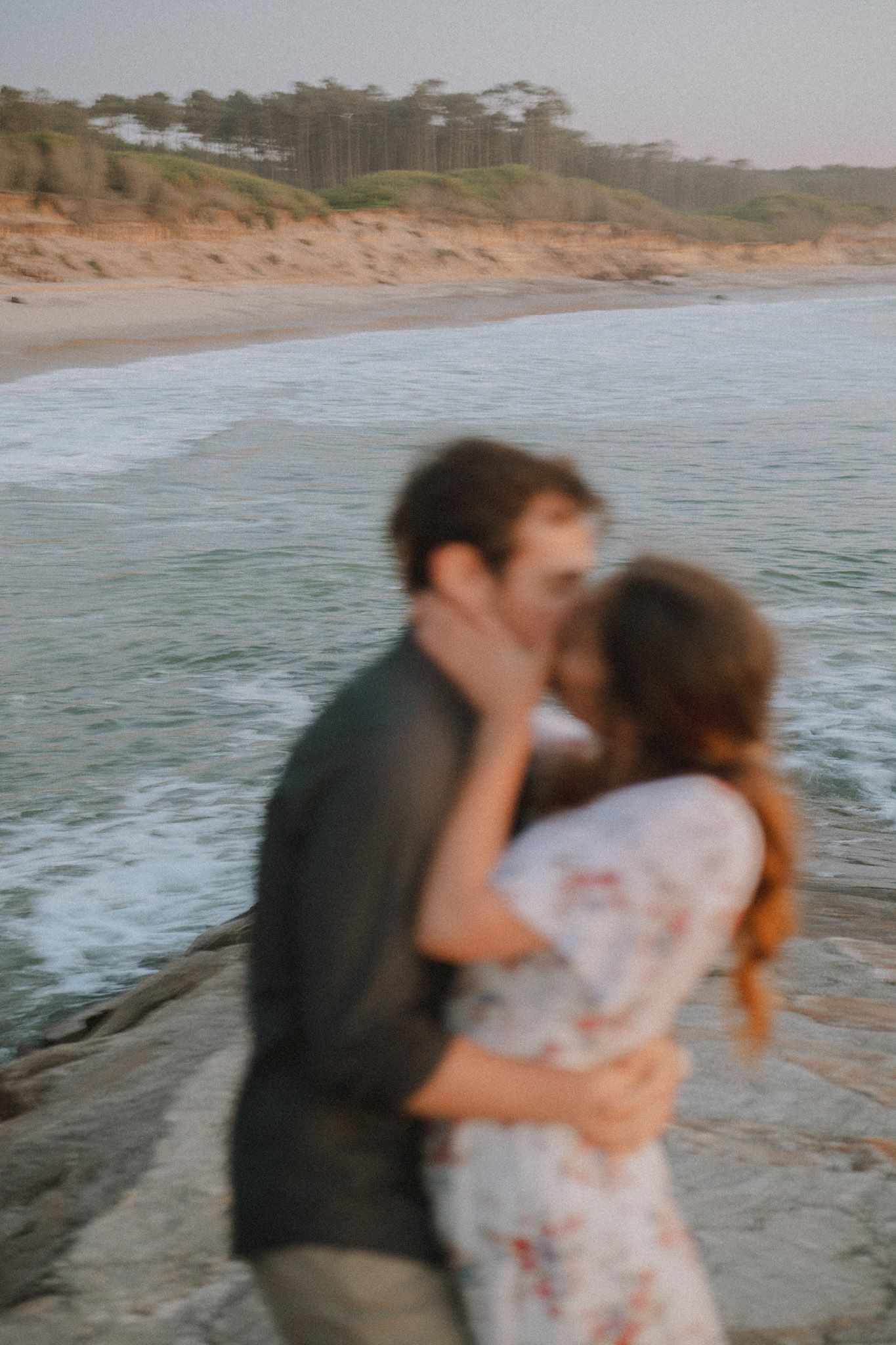 Couple embracing on rocky coastline with ocean waves in background at sunset.