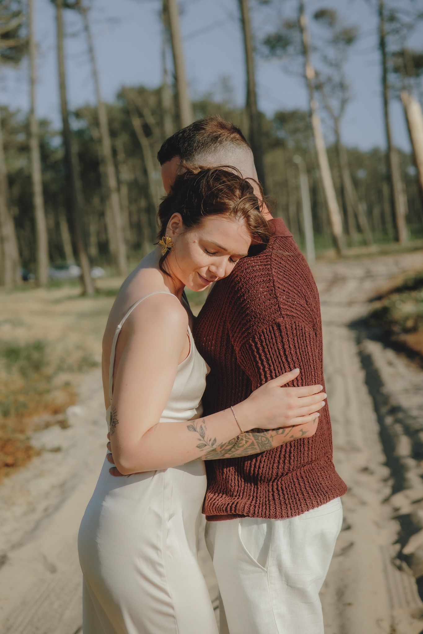 Intimate moment captured between two people in white and burgundy outfits against a coastal backdrop.