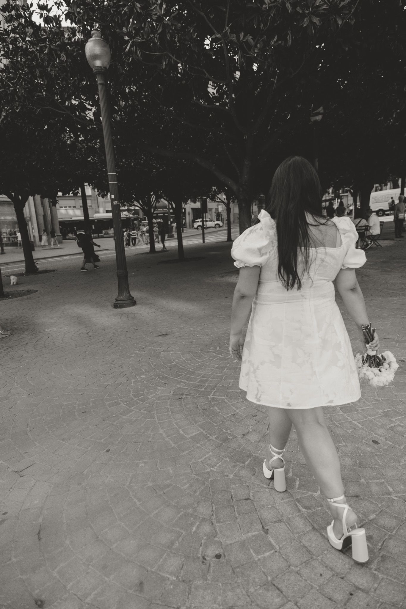 Black and white photo of a flowing white dress and bouquet moving across a brick paved path in a park setting.