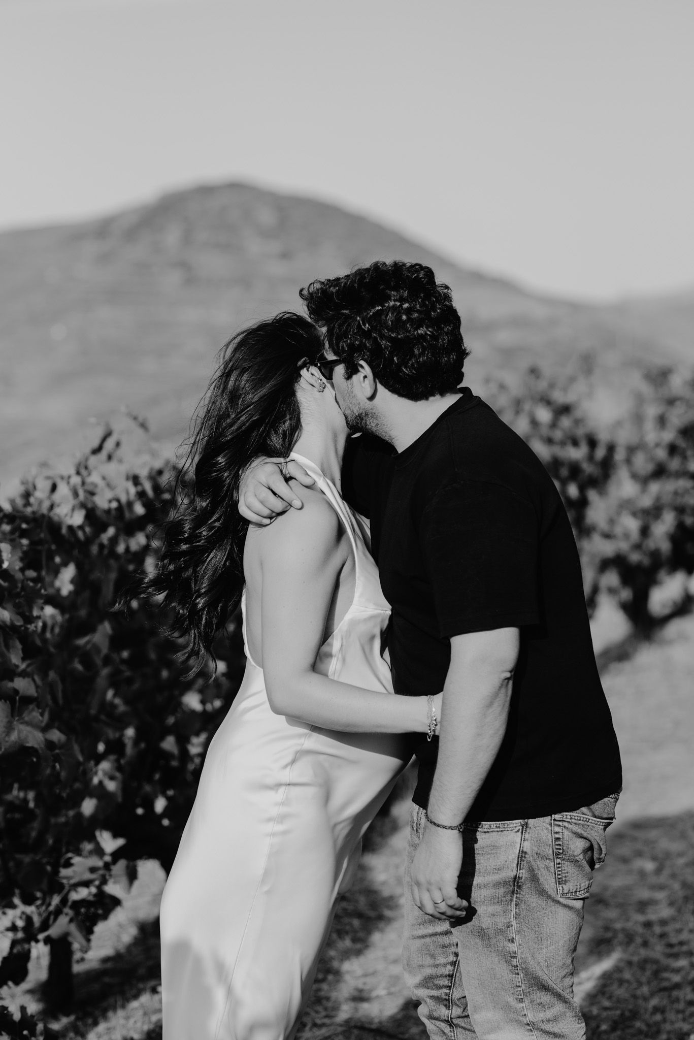 A romantic couple shares intimate kisses in a vineyard with rolling hills in the background in black and white photography.