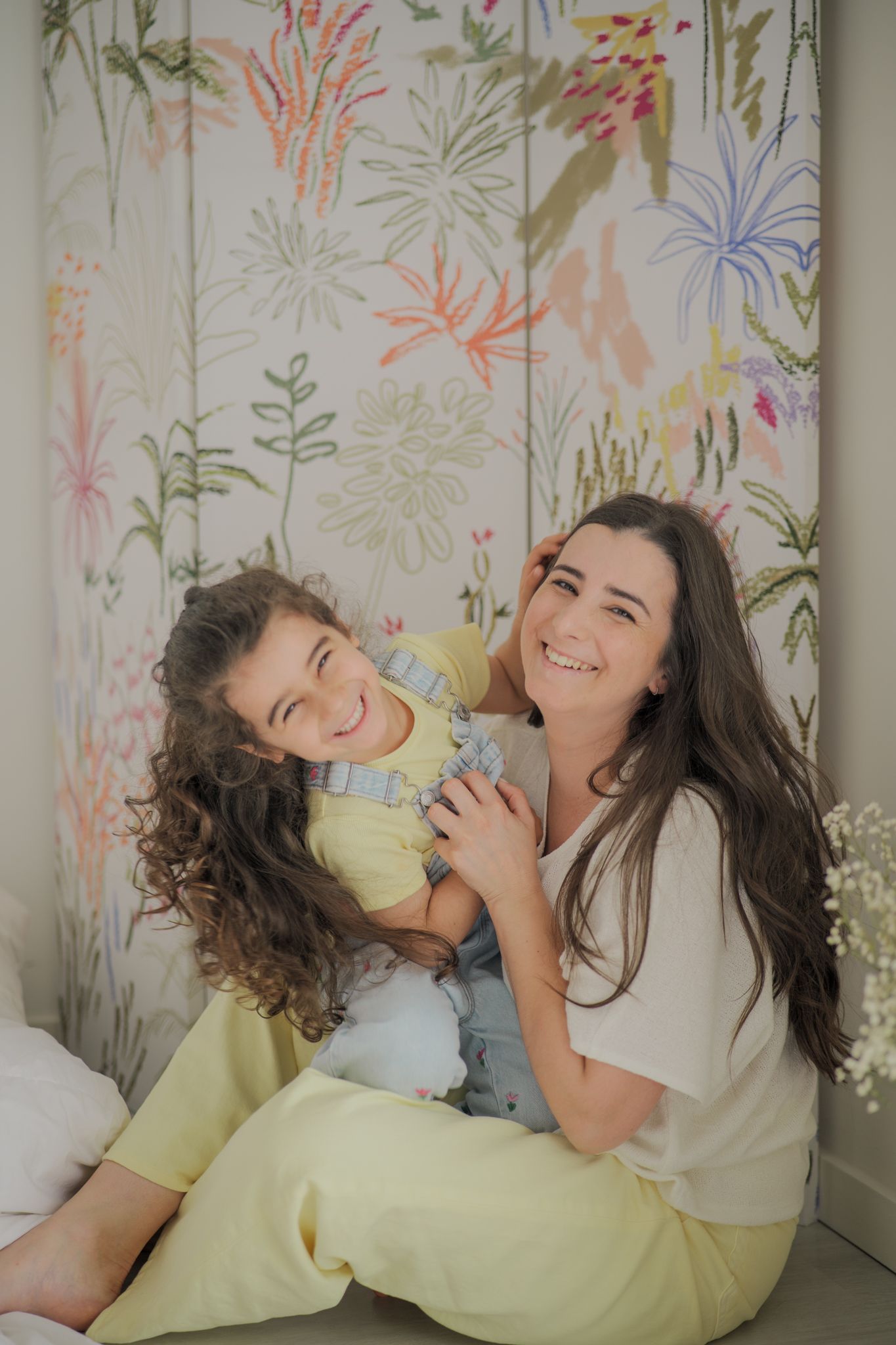 A smiling mother and daughter sitting together in front of a colorful floral wallpaper, wearing yellow outfits.