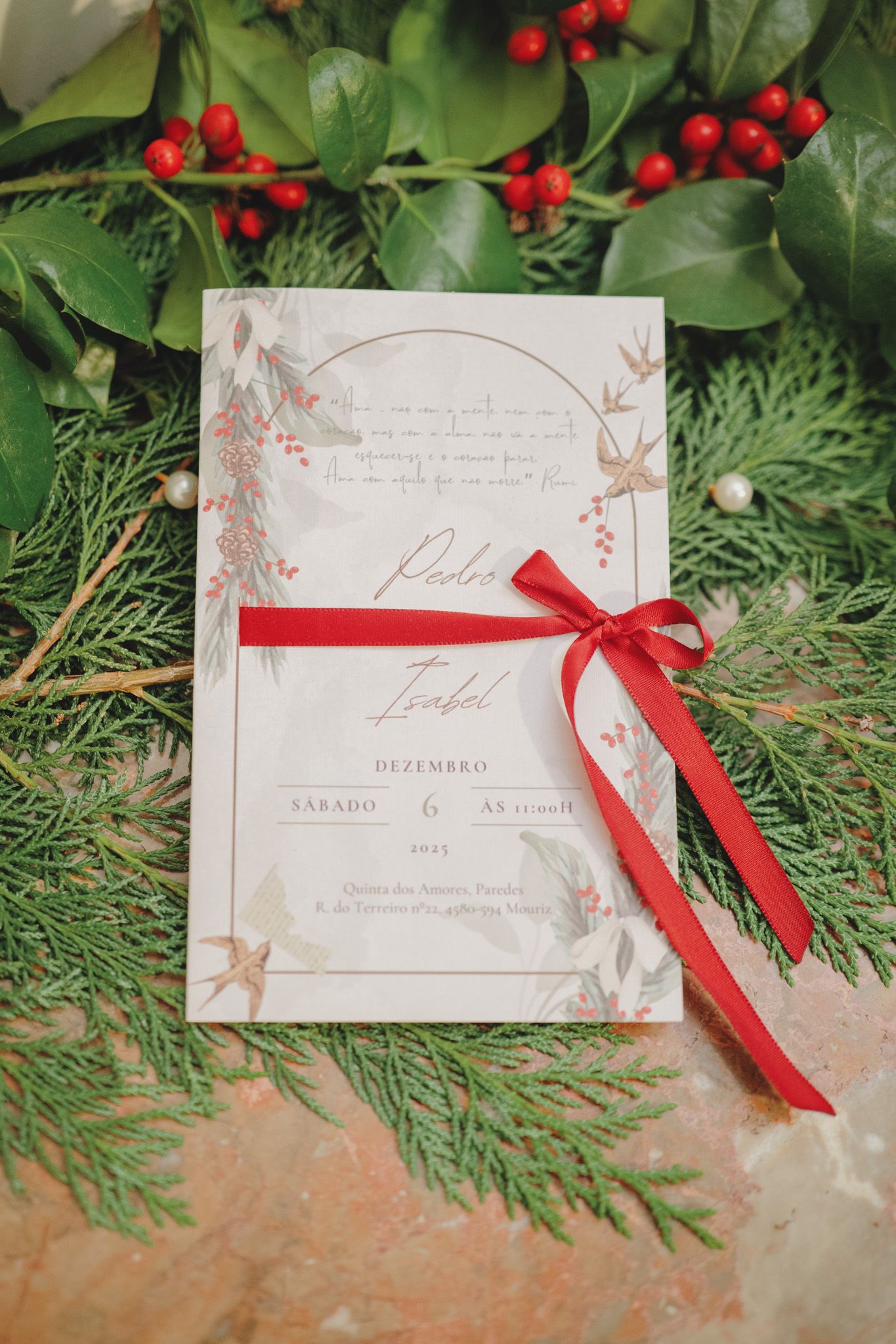 Wedding invitation with red ribbon surrounded by holly berries and evergreen branches.