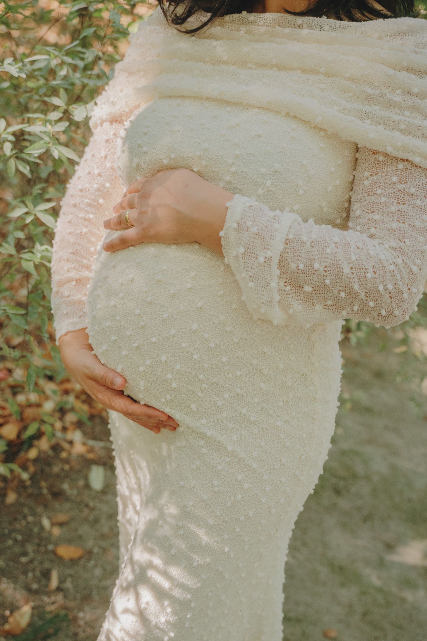 Close-up of a white pearl-embellished maternity dress showing pregnant belly in an outdoor garden setting.