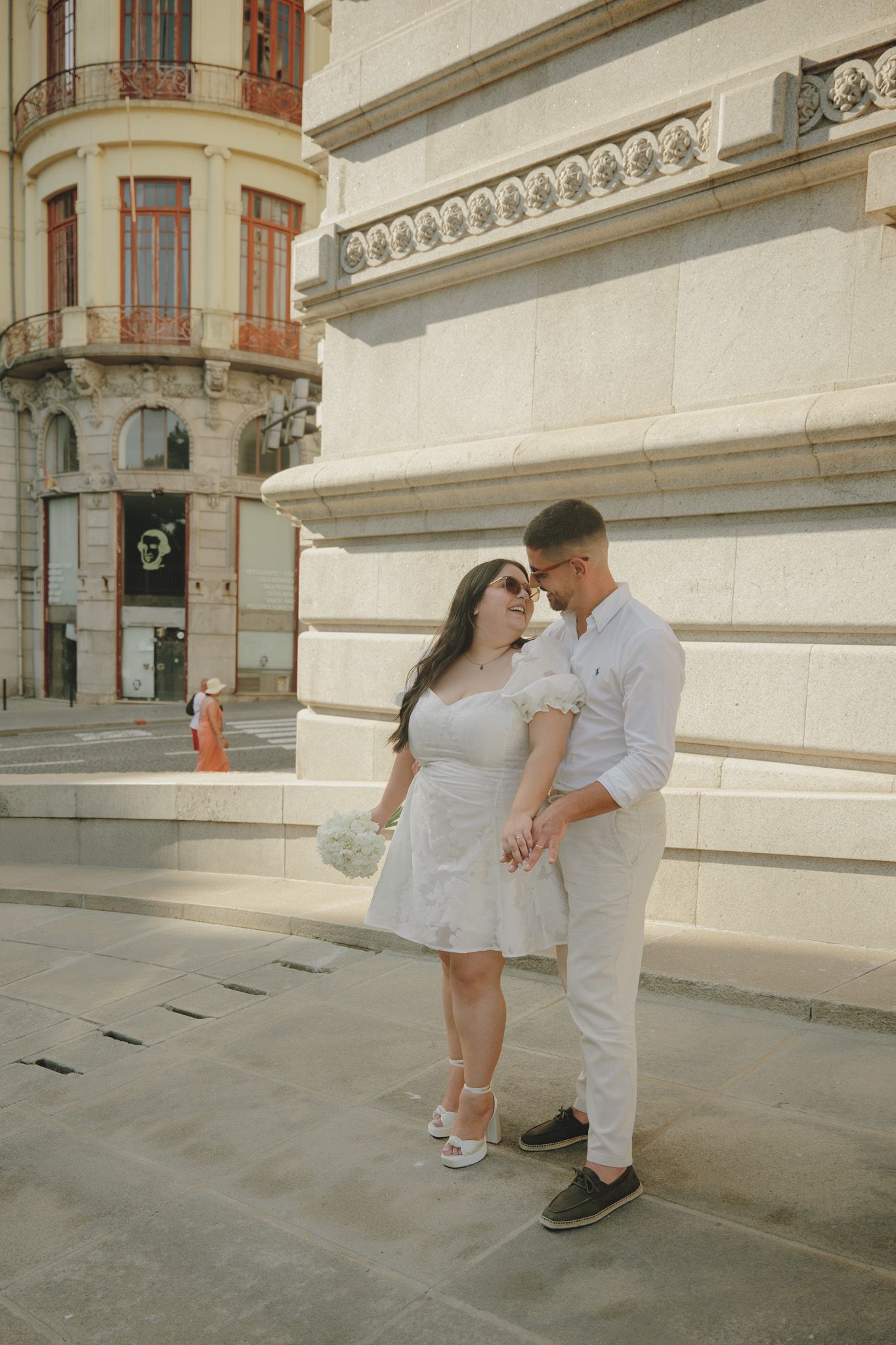 A couple in white outfits shares intimate moments against a classic building facade.