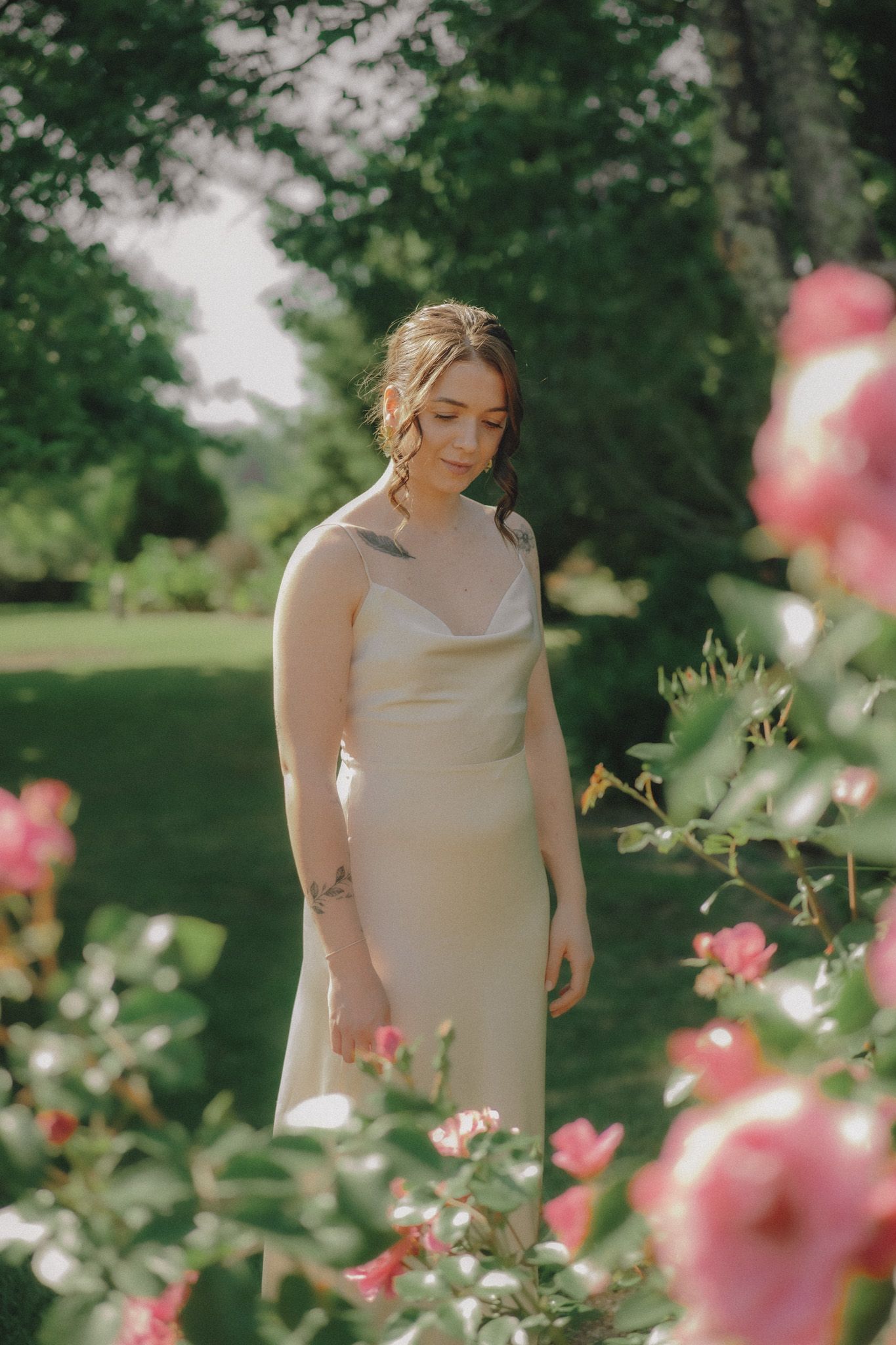 Bride in silk dress standing among blooming pink roses in garden setting.