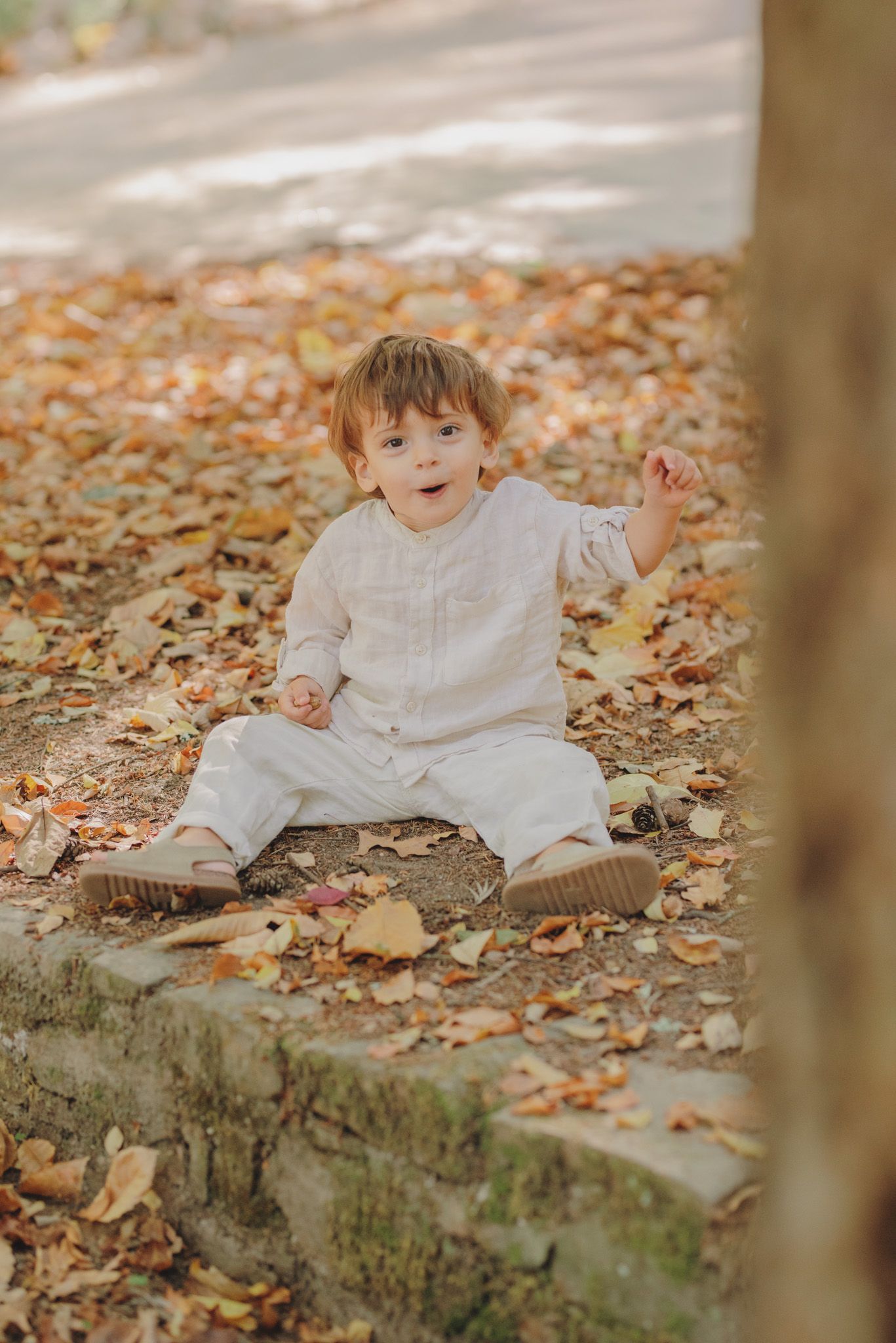 A joyful toddler in white clothing sits on stone steps covered with autumn leaves.