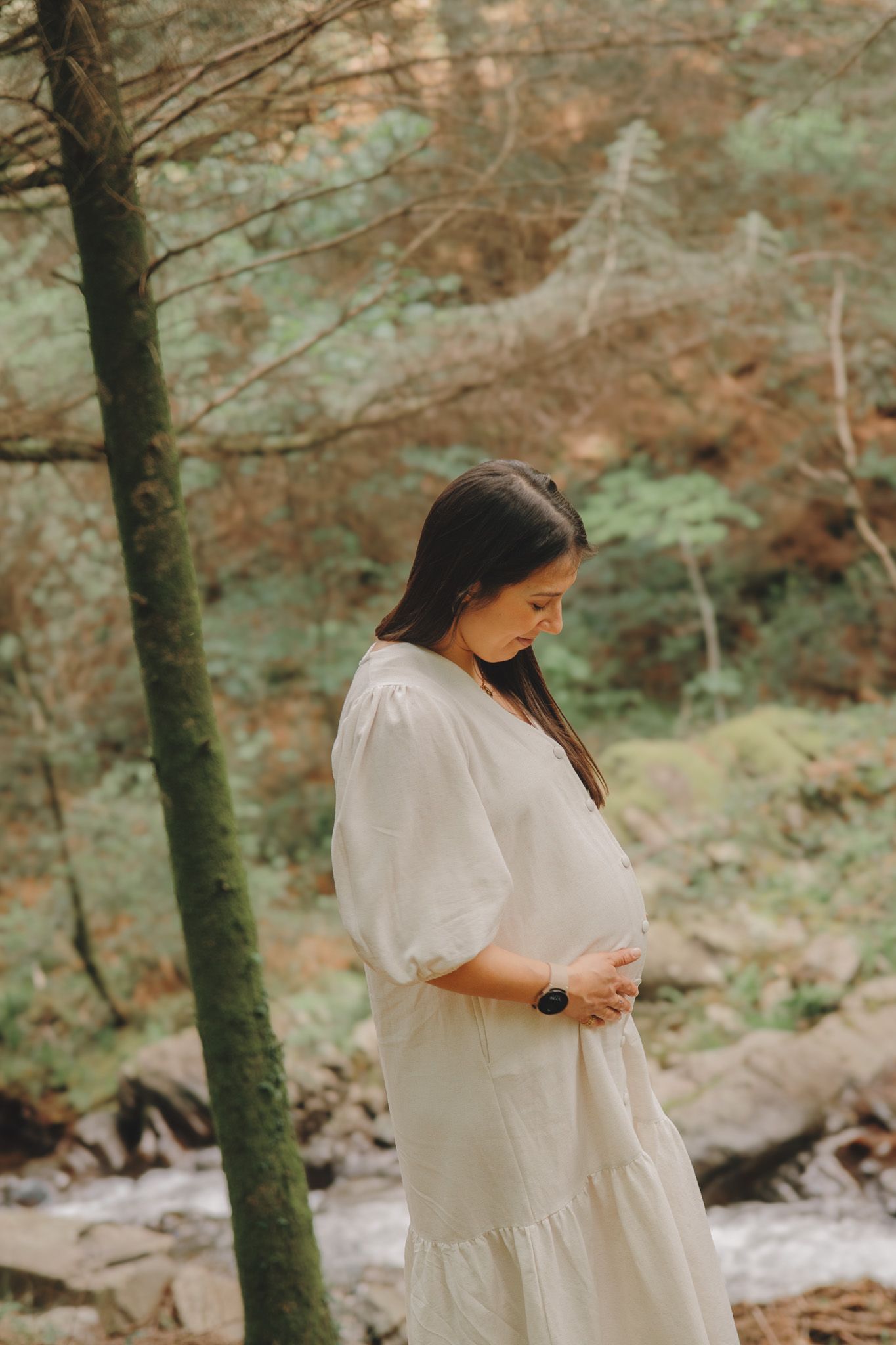 Figure in white dress stands on forest trail beside a flowing stream.