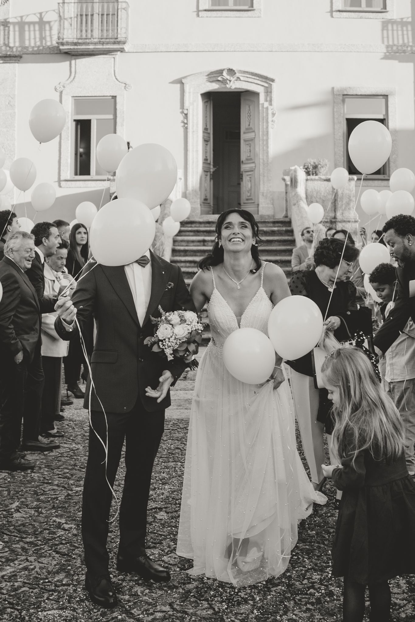 Black and white photo of wedding guests gathered with white balloons outside historic building.