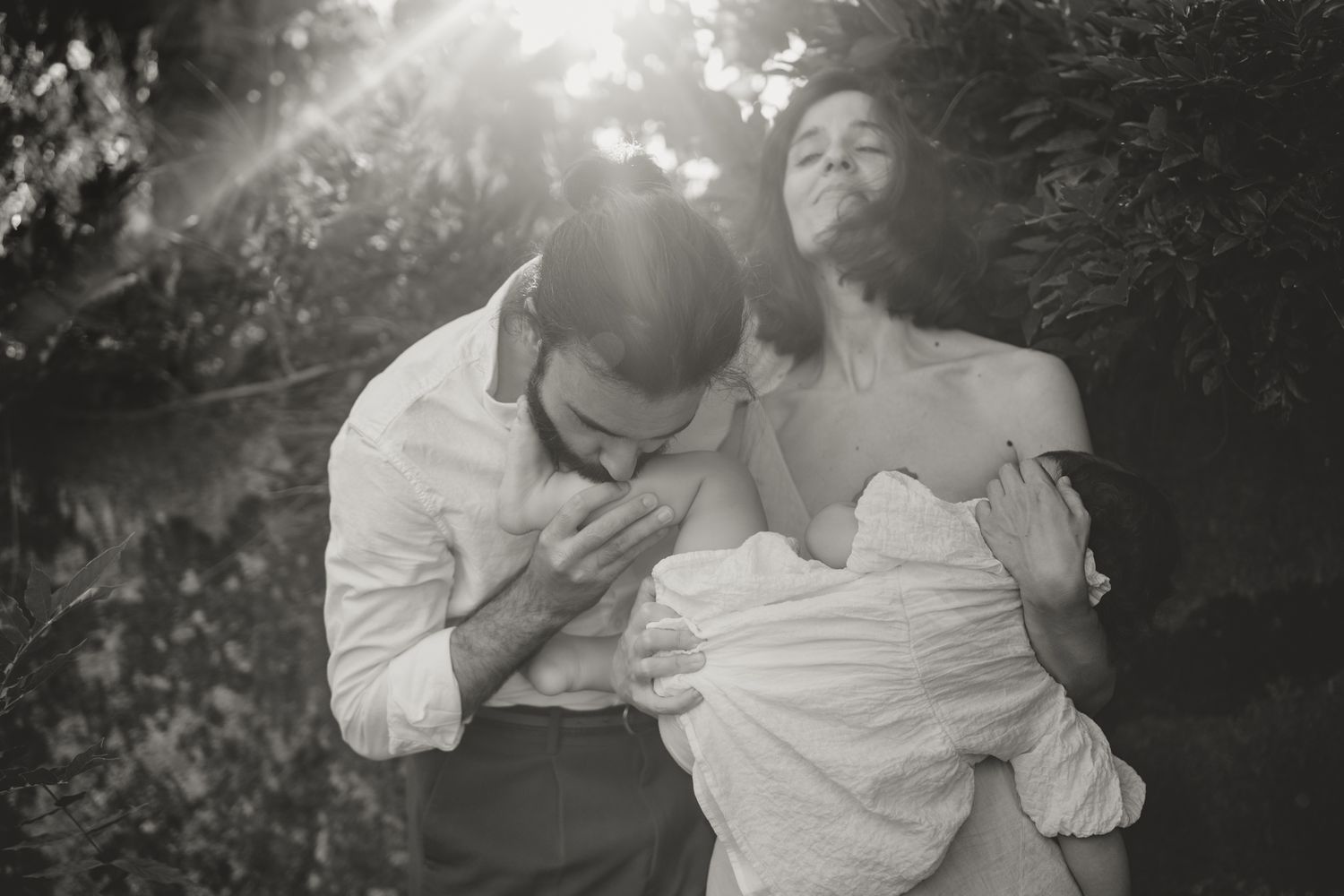 A romantic black and white portrait of a couple embracing outdoors with sunlight streaming through trees.