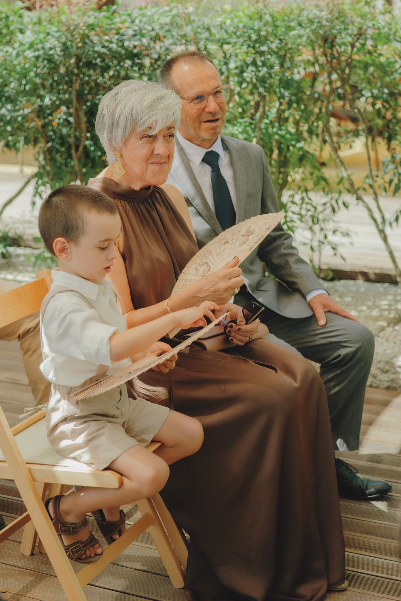 A young child sits with family members on wooden chairs during an outdoor gathering.