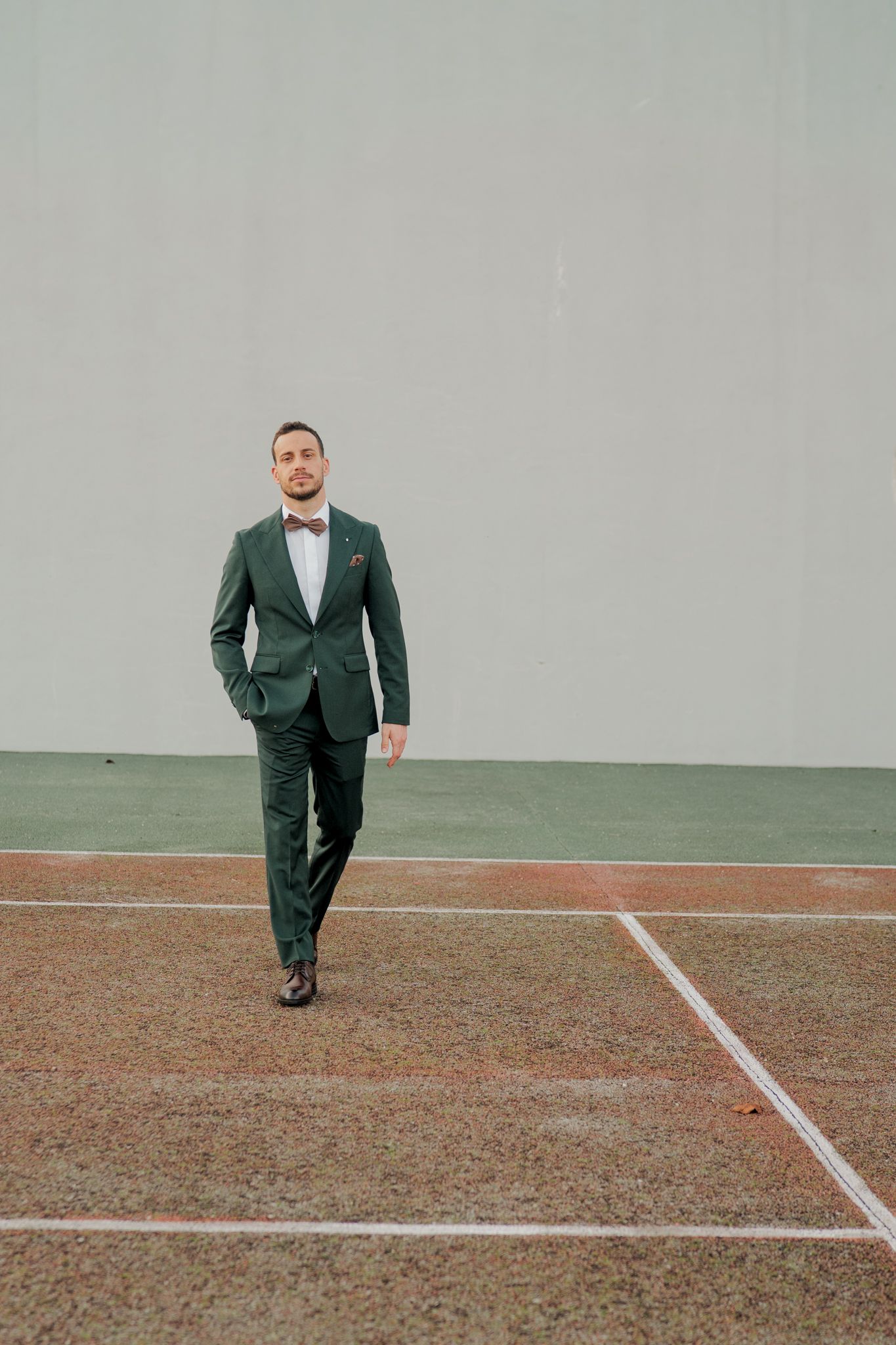 Groom in emerald green suit walks across sports court during creative wedding photoshoot.